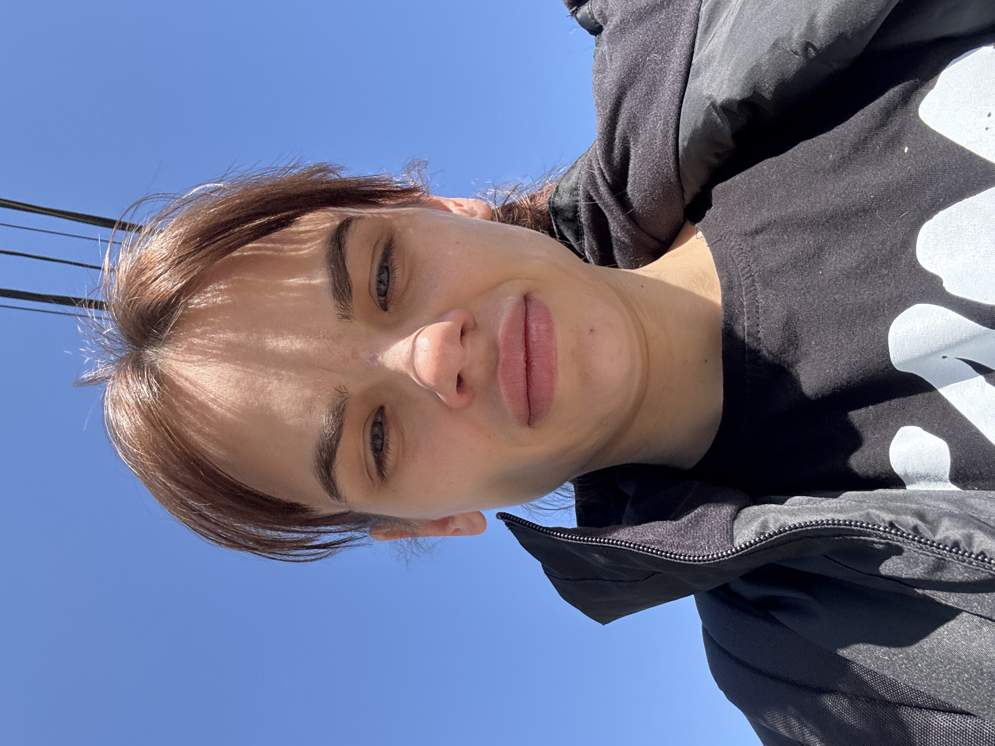 The image shows a selfie taken from a low angle, featuring a person with fair skin, brown hair, and light eyes against a clear blue sky. The person is wearing a black hooded jacket with a black t-shirt underneath that has a white design. In the upper left corner of the frame, there are black wires. The absence of specific landmarks or indicators of Rostov-on-Don makes it impossible to confirm the location.