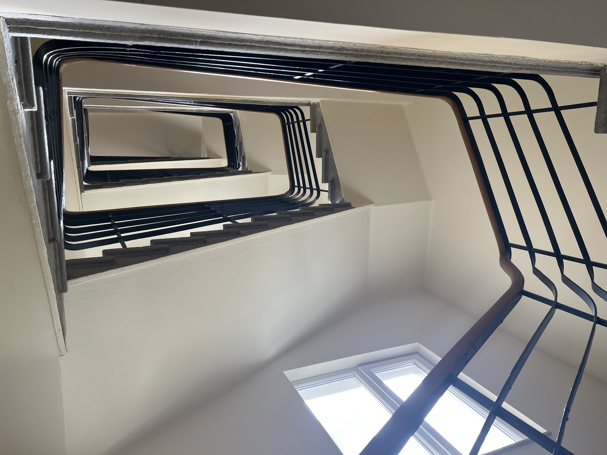 A low-angle shot of a spiral staircase is presented. The staircase has a square opening in the center, with multiple levels visible, receding upwards. The stair treads appear light gray. The walls are painted white. A square window is located at the bottom of the frame. The staircase is framed by a black metal railing that curves along the stairs. The lighting is bright, illuminating the stairwell.
The location in the city of Sofia, Bulgaria cannot be determined based on provided information.