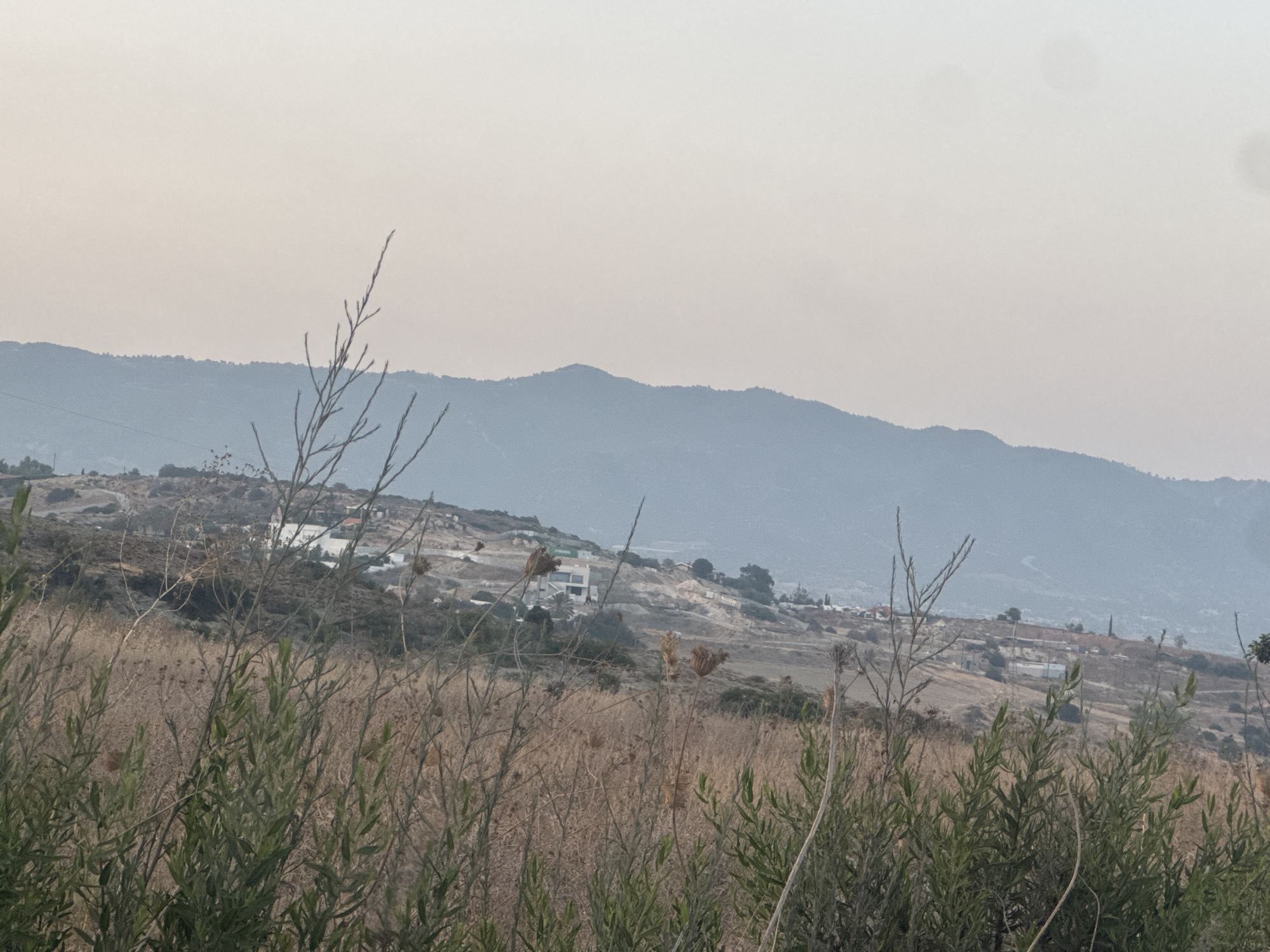 The image depicts a hazy, mountainous landscape near Agios Tychona Community, Cyprus, on a clear day. In the foreground, dry grasses and green shrubbery are prominent, with a few bare branches extending upwards. The middle ground reveals a hillside dotted with buildings and some construction activity. The background is dominated by undulating hills and mountains fading into the pale sky. The overall scene conveys a natural, somewhat arid environment with a hint of human presence.