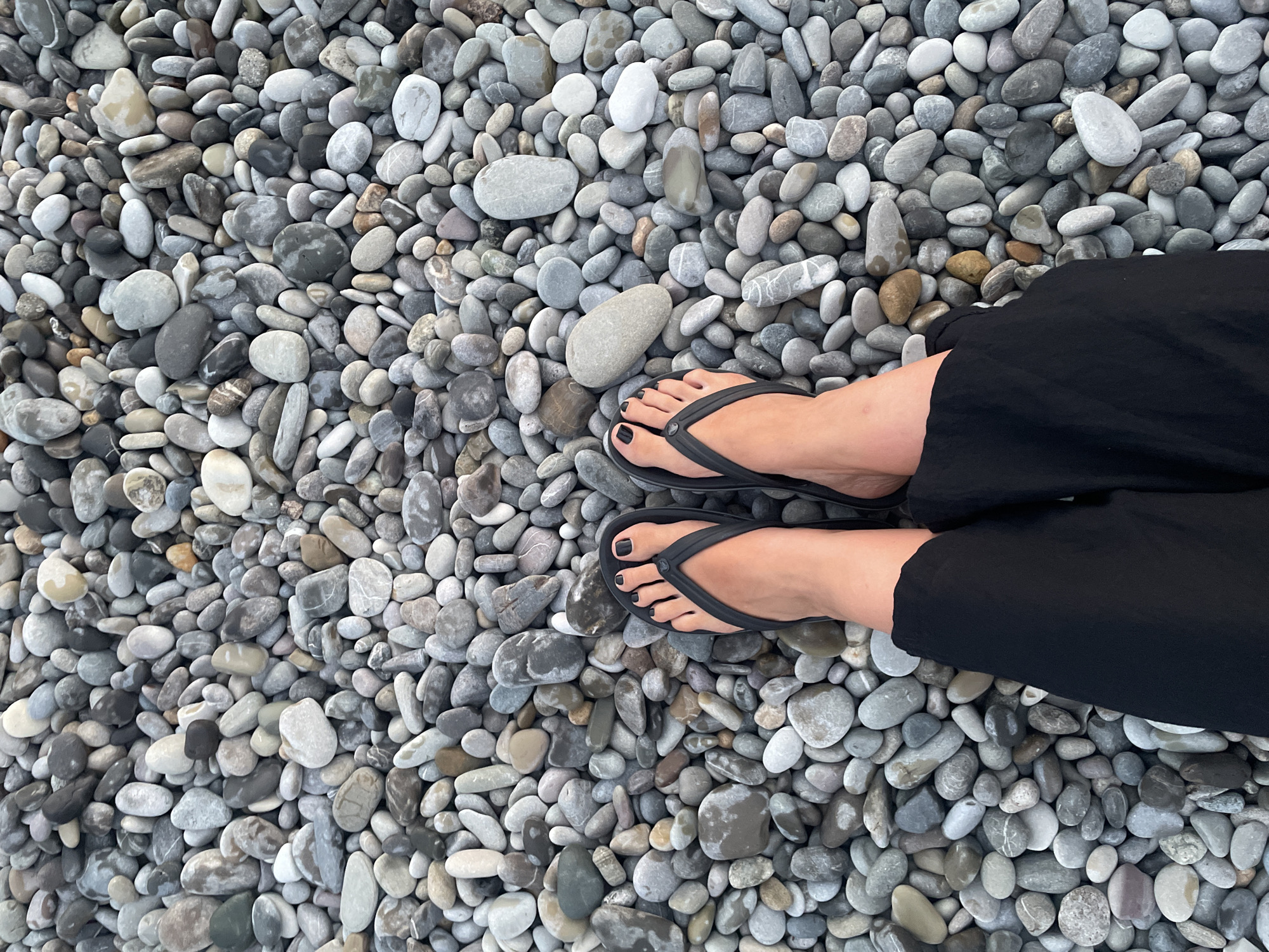 A top-down, medium shot captures a pair of feet, shod in black flip-flops, resting on a dense bed of smooth, gray and white pebbles. The feet are clean, with black-painted toenails. The individual is wearing black pants that extend below the knees. The pebbles are varied in size and shape, with some appearing wet and others dry, suggesting a beach or shoreline environment. The lighting is even, indicating an overcast day or ambient light conditions. The background consists entirely of the pebble-covered ground.