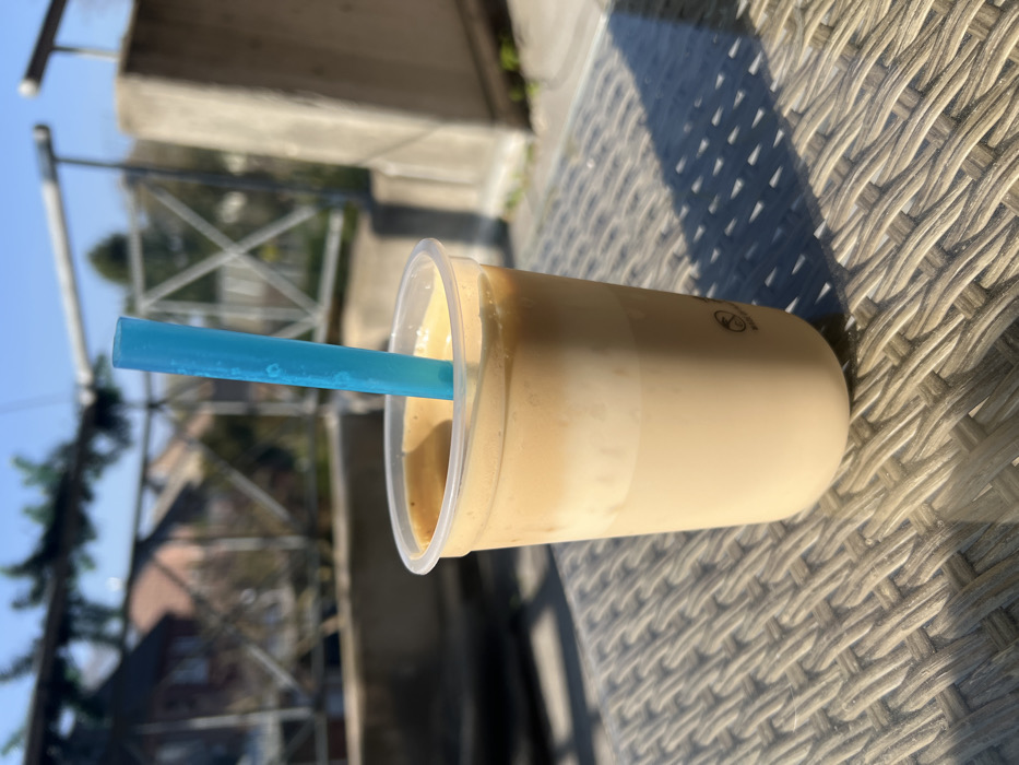 A clear plastic cup containing a layered iced beverage, approximately two-thirds full, rests on a light-colored, woven synthetic rattan table. The beverage consists of a lighter, opaque lower layer and a darker, frothy upper layer. A light blue, translucent cylindrical straw is inserted into the drink. Strong, direct sunlight illuminates the scene, casting distinct shadows from the cup and the table's weave onto the table surface.
In the background, out of focus, a metal railing system with horizontal bars and vertical supports is visible, partially obscured on the left by green foliage. Beyond the railing, several residential buildings with reddish-brown pitched roofs are discernible under a clear blue sky. A concrete support pillar forms part of the railing structure on the right. The scene depicts an outdoor setting, likely a balcony or terrace, in the city of Würselen, Germany. No individuals or specific actions are present.