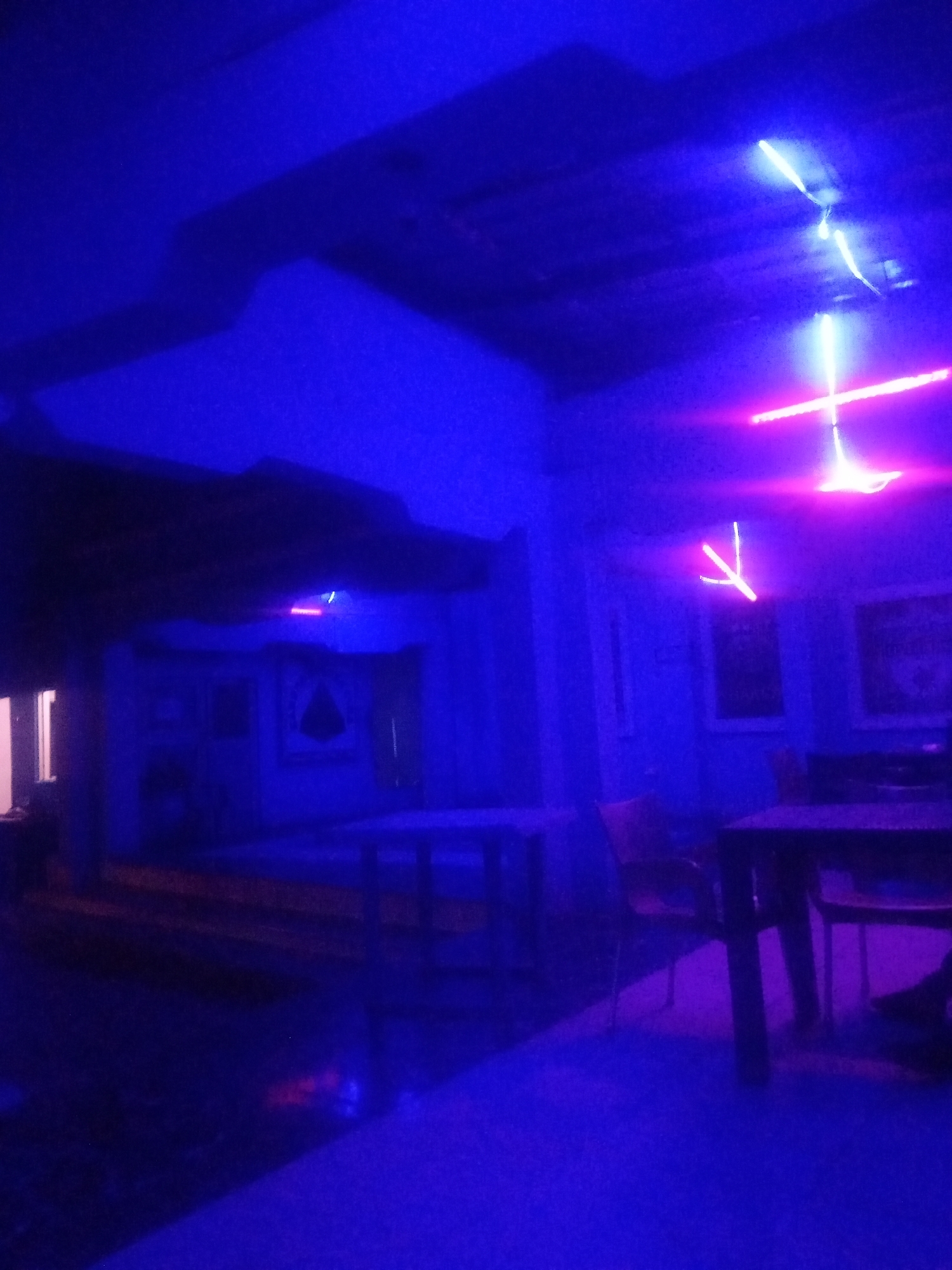 The image depicts an interior scene illuminated by vibrant blue and pink neon lights. The setting appears to be a dimly lit establishment, possibly a bar or lounge, with furniture including tables and chairs visible. The lighting creates a dramatic and somewhat abstract atmosphere, with shadows and light reflections playing across the surfaces. There are no discernible individuals clearly visible, though the presence of furniture suggests it is a public space. The environment is characterized by a modern, possibly trendy, aesthetic due to the lighting choices. The location specified, Sabon Gari Nassarawa, Nigeria, provides a geographical context for this urban interior.