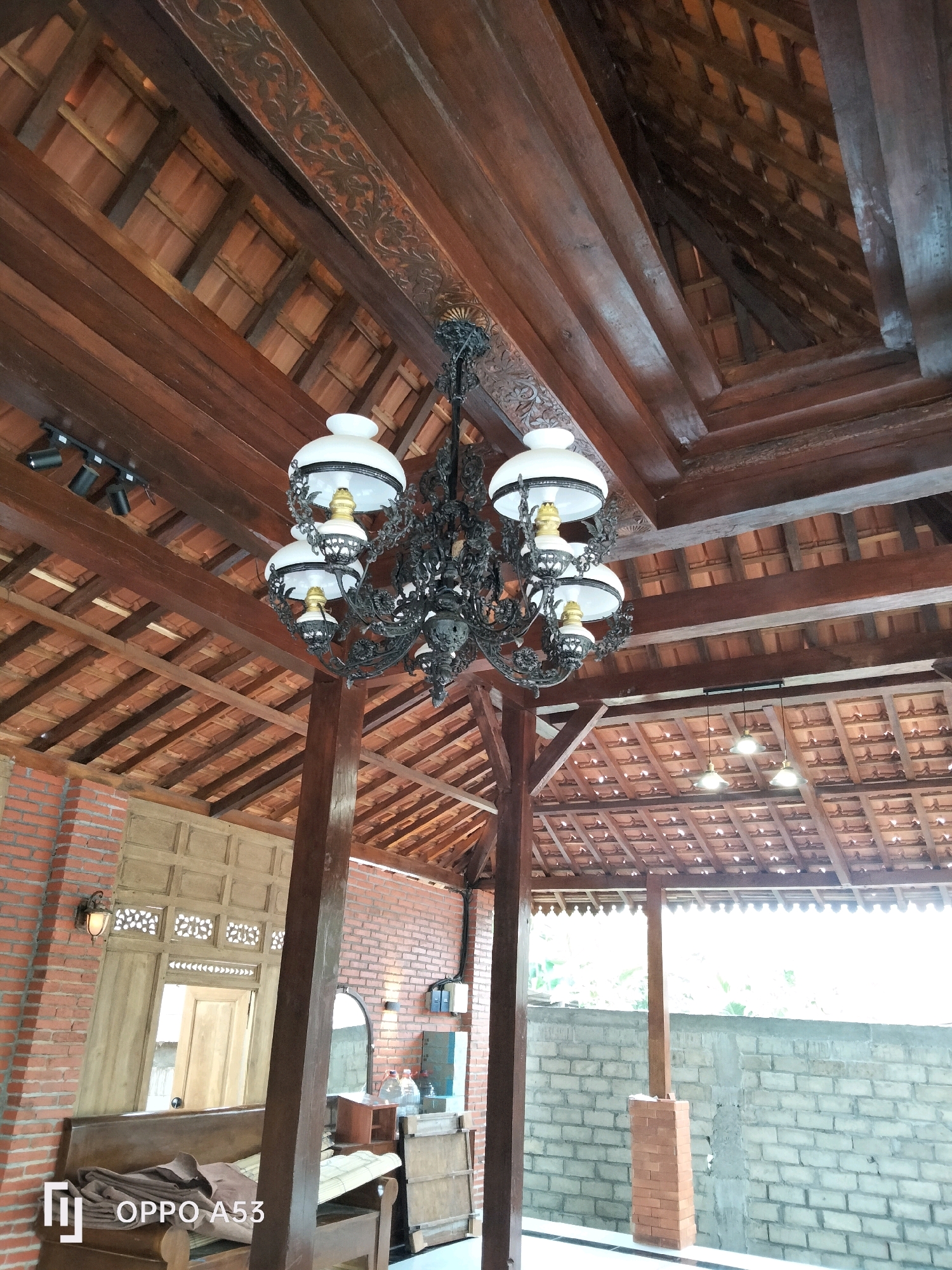 The image depicts an interior scene of a building with a traditional Indonesian architectural style, likely a home or a restaurant, located in Trenggalek, Indonesia. The ceiling is constructed with exposed wooden beams and red clay tiles, creating a warm and rustic ambiance. A large, ornate black chandelier with white lamps hangs from one of the main beams, serving as a prominent focal point. Additional lighting fixtures, including track lighting and smaller pendant lights, are also visible on the ceiling. The walls are made of red brick and wooden panels, further enhancing the traditional aesthetic. In the foreground, a wooden bench with a folded blanket is partially visible. Other objects in the scene include a mirror, bottles of water, and what appears to be a cabinet or dresser. The overall impression is one of a well-preserved traditional Indonesian interior, possibly a cultural or heritage space within the city of Trenggalek.