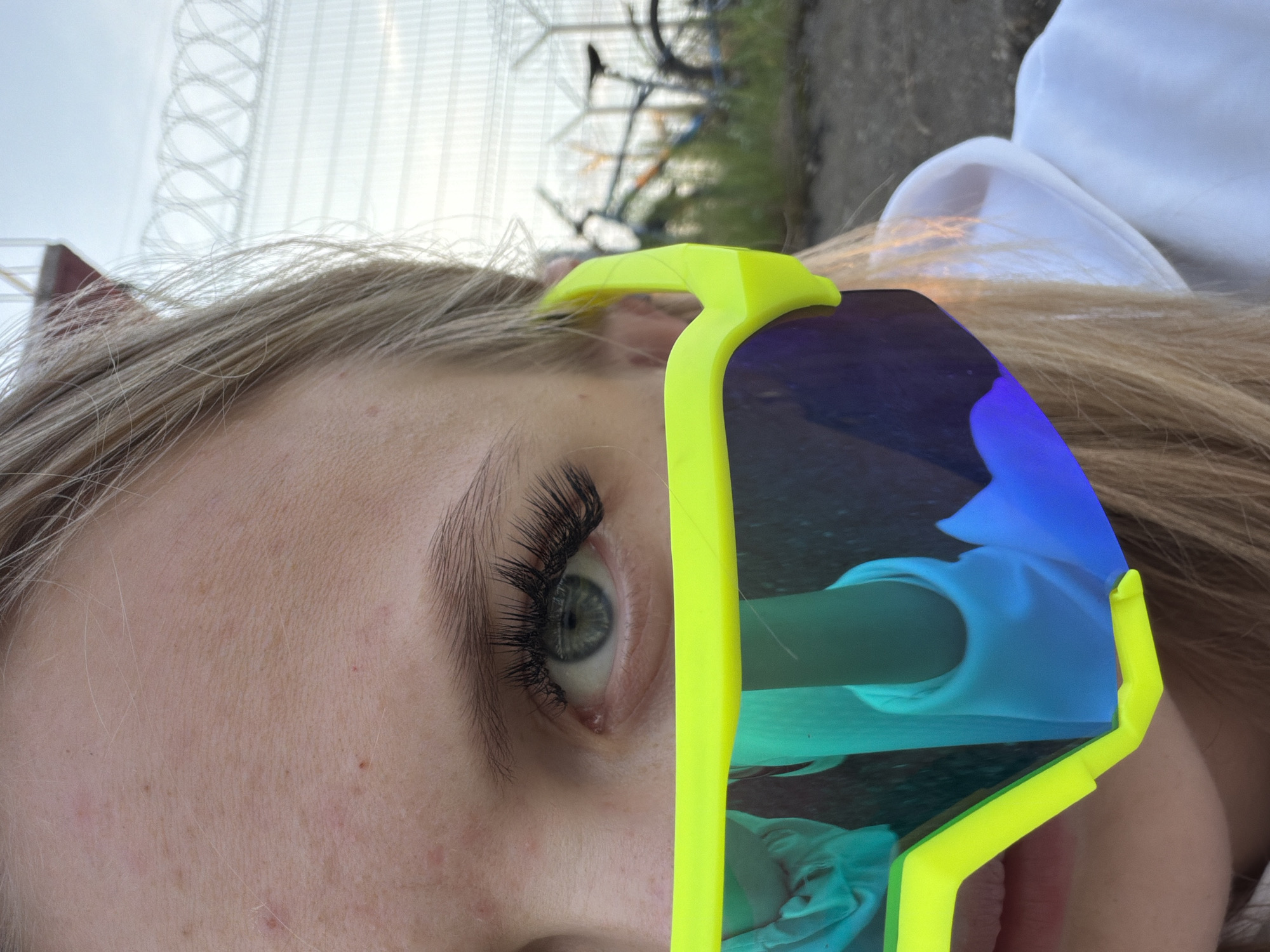 A close-up shot features the right side of a young woman's face, including her eye, eyebrow, and partial forehead and cheek. She is wearing large, neon yellow sports goggles with mirrored lenses that reflect a blue and teal sky, along with a blurry teal sleeve. Her blonde hair is visible on the right side of the frame. In the background, a fence with barbed wire runs horizontally, and two bicycles are parked on a dirt path with scattered grass. The lighting suggests it is daytime, possibly late afternoon or early morning, with the sun low on the horizon. The overall setting appears to be an outdoor area with an industrial or security-related structure in the distance. The image is not specific enough to determine its precise location within the city of Chaikovsky, Russia, but it could be an area on the outskirts or near a facility.