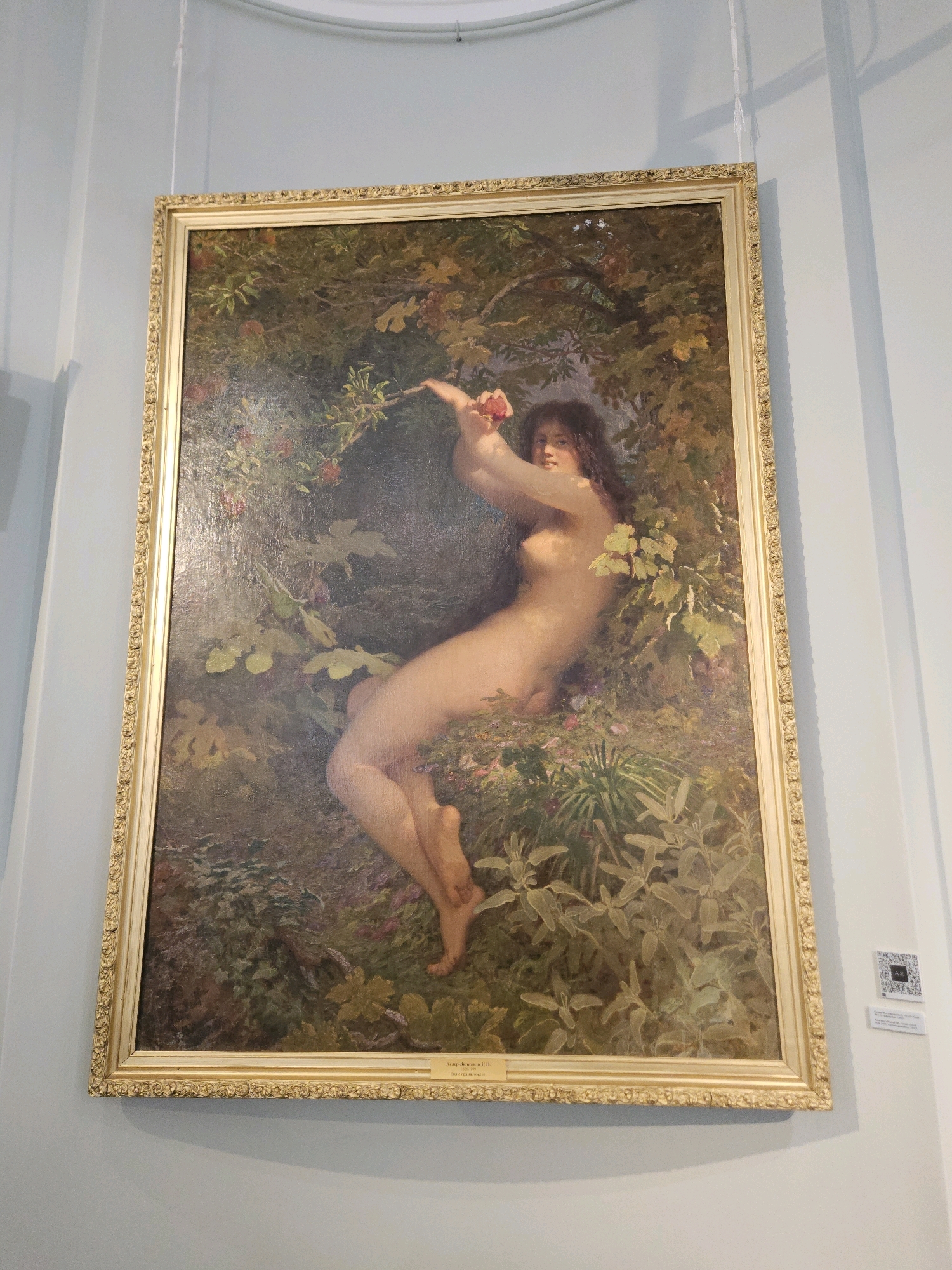 This is a painting hanging on a pale blue wall, framed in ornate gold. The painting depicts a nude woman with long, dark hair, reclining amongst lush foliage and red fruits, possibly apples or berries. She is reaching up with her right hand towards a small, red fruit, and her left arm is bent, with her hand resting on her chest. The woman's body is pale and appears to be in a natural, relaxed pose. The background is filled with various shades of green and brown leaves and branches, with some hints of purple flowers and more red fruits. The lighting suggests an outdoor, natural setting, possibly a garden or orchard. A small white label with text is visible on the bottom edge of the painting's frame, and a QR code and another label are affixed to the wall to the right of the painting. The location is the Ulyanovsk Regional Art Museum, which houses works by the artist I. N. Klyushin, as indicated by the label beneath the painting.