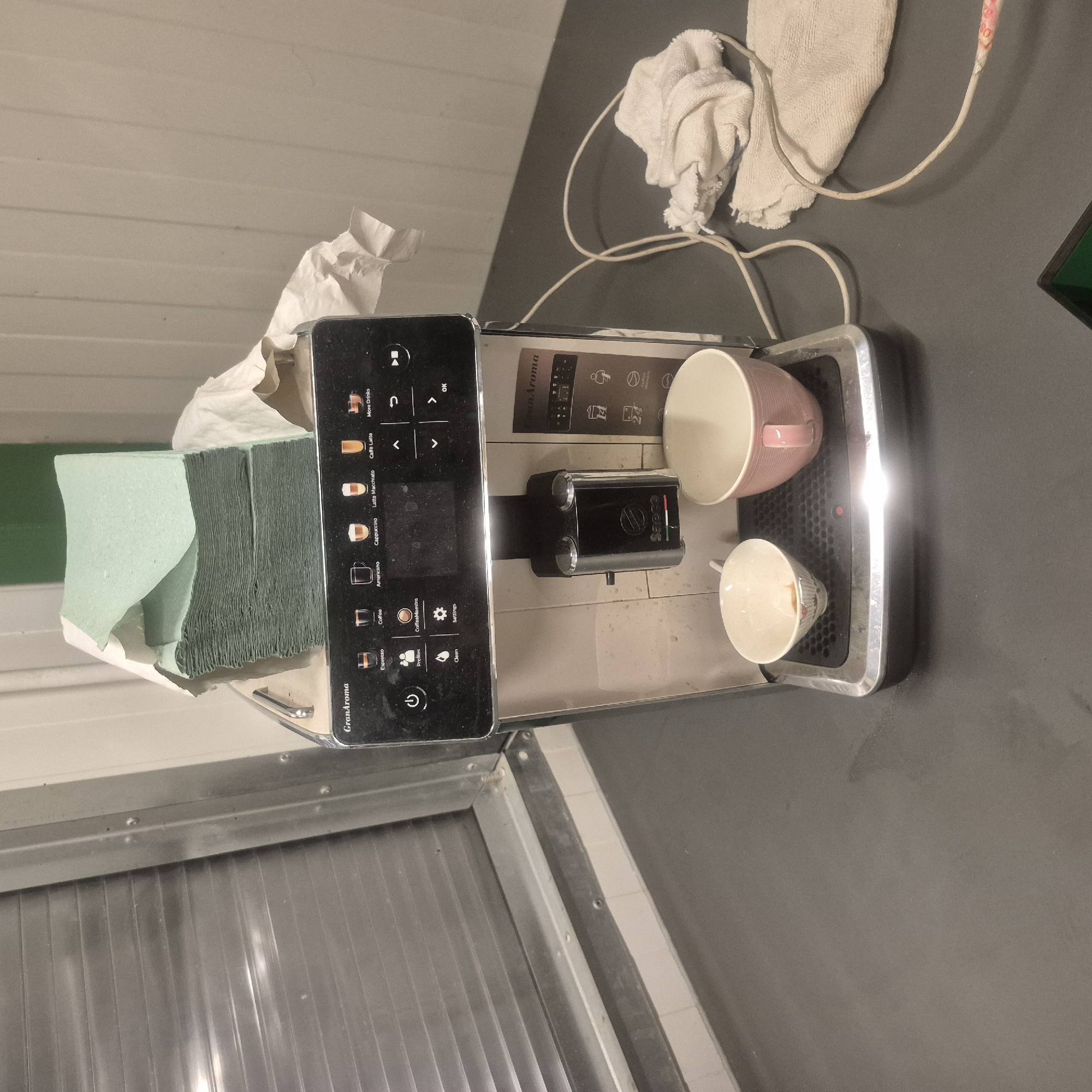 A Saeco GranAroma automatic coffee machine sits on a dark gray countertop in what appears to be a utility room or storage area. The machine is silver and black with a touch screen display showing various coffee options and controls. A stack of folded green paper napkins is piled on top of the machine, partially covered by a white cloth. Two cups are positioned on the drip tray of the coffee machine; one is a small white cup with a floral pattern, and the other is a larger pink ceramic mug. A white power cord snakes across the countertop, and a crumpled white cloth lies near the coffee machine. In the background, a large silver slatted blind covers a window or door. The walls are a neutral, light color. The overall impression is one of a functional, somewhat cluttered space, possibly a break room or kitchen area. The scene does not provide any specific indicators of location within Nowa Wieś Książęca, Poland, beyond being a potential interior scene in that region.
