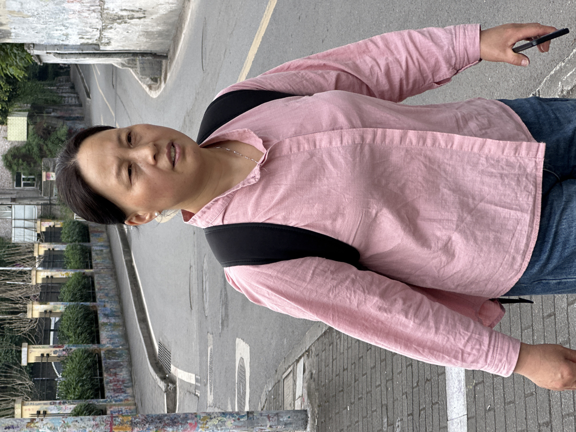 A woman in a pink long-sleeved shirt, blue jeans, and a black backpack is standing on a gray sidewalk in Jiulongpo District, China. She is holding a black cell phone in her right hand. The background features a gray paved street with a yellow dividing line, buildings with greenery, and a decorative fence.
