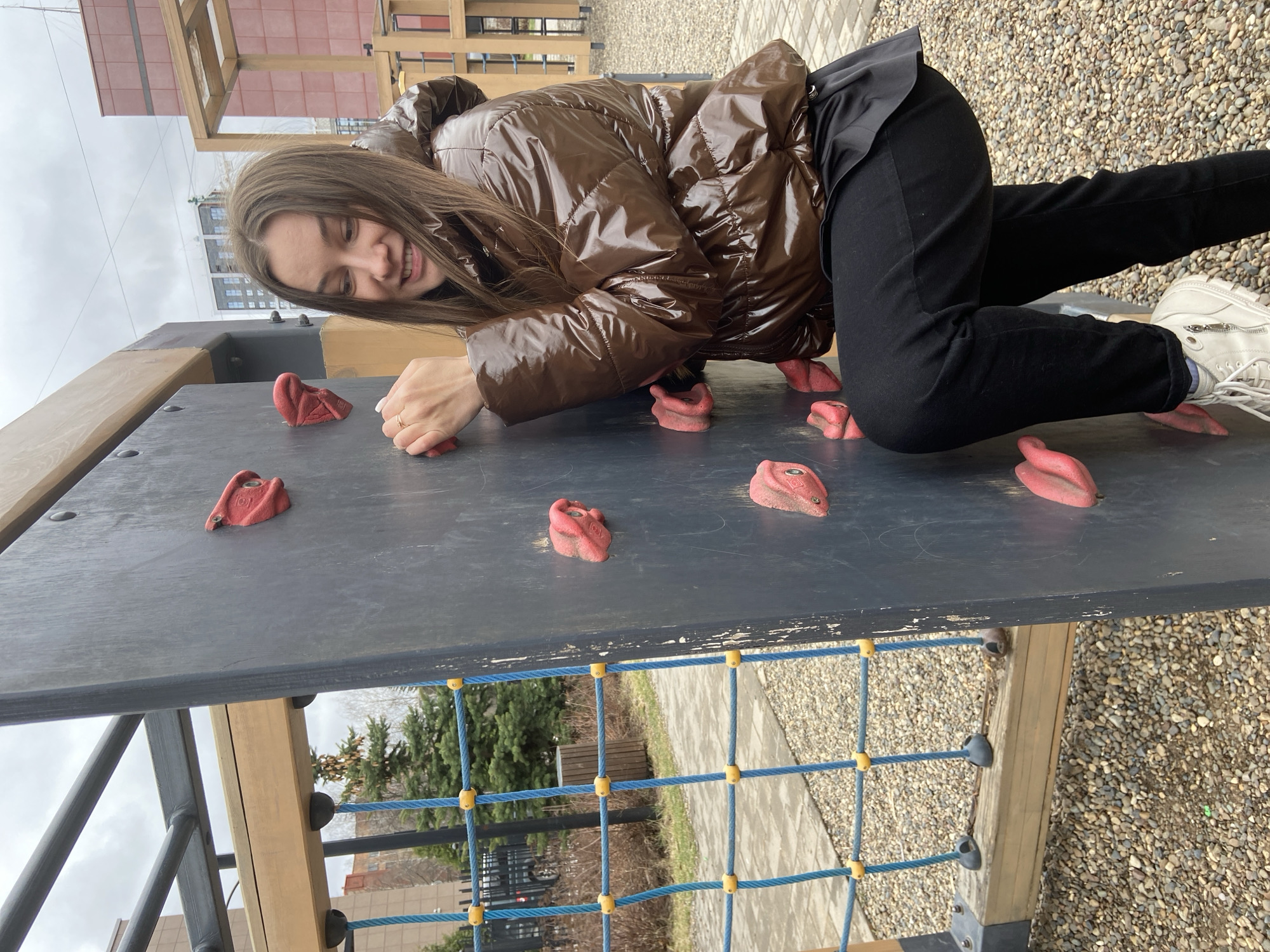 A young woman is playing on a climbing wall in a playground. She is wearing a brown puffer jacket and black pants, and is using the red climbing holds to support herself. The climbing wall is part of a larger play structure with wooden beams and a rope net. The ground is covered in gravel. In the background, there are trees and buildings, including a high-rise. Based on the style of architecture, language and imagery, it appears the photo may have been taken in the city of Izhevsk, Russia.