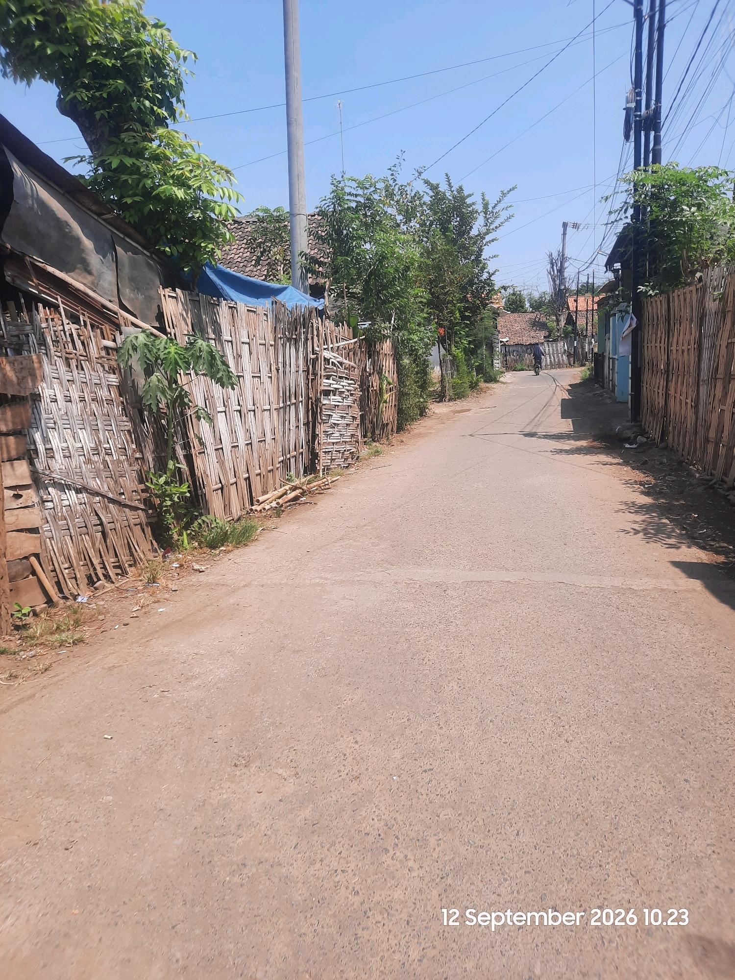 The image depicts a rural road in Sentong, Indonesia, on September 12, 2026, at 10:23 AM. The road is made of asphalt and is lined with bamboo fences and vegetation on both sides. A person is riding a motorcycle in the distance, heading away from the camera. Overhead power lines crisscross the sky, supported by utility poles. The architecture visible in the background suggests a residential area with houses featuring tiled roofs. The weather appears to be clear and sunny.