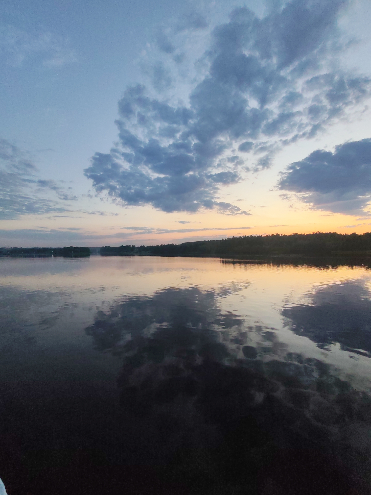 The image depicts a serene waterscape in Ermolino, Russia, likely at dusk or dawn. The sky transitions from a pale blue to a soft orange near the horizon, with numerous gray and white clouds scattered across the upper portion of the frame. Below the horizon line, a dense treeline is visible. The water, which appears calm, reflects the sky and clouds, creating a mirror-like effect. The foreground gradually darkens into the depths of the water, possibly obscuring the shoreline. The scene is devoid of people or other distinct subjects, focusing primarily on the natural elements.