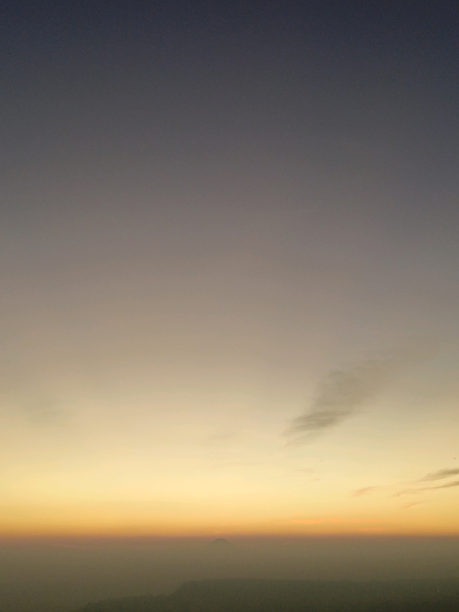 This image is a landscape photograph of the sky at dawn or dusk, with a gradient of colors from deep blue at the top to bright orange and yellow at the horizon. A faint silhouette of mountains or hills is visible in the distance, partially obscured by a haze. There are a few wispy clouds in the sky. The lower portion of the image shows a dark, indistinct landscape. The scene is devoid of any human presence or specific man-made structures that would allow for definitive identification of a precise location within Butuh Kidul, Indonesia.