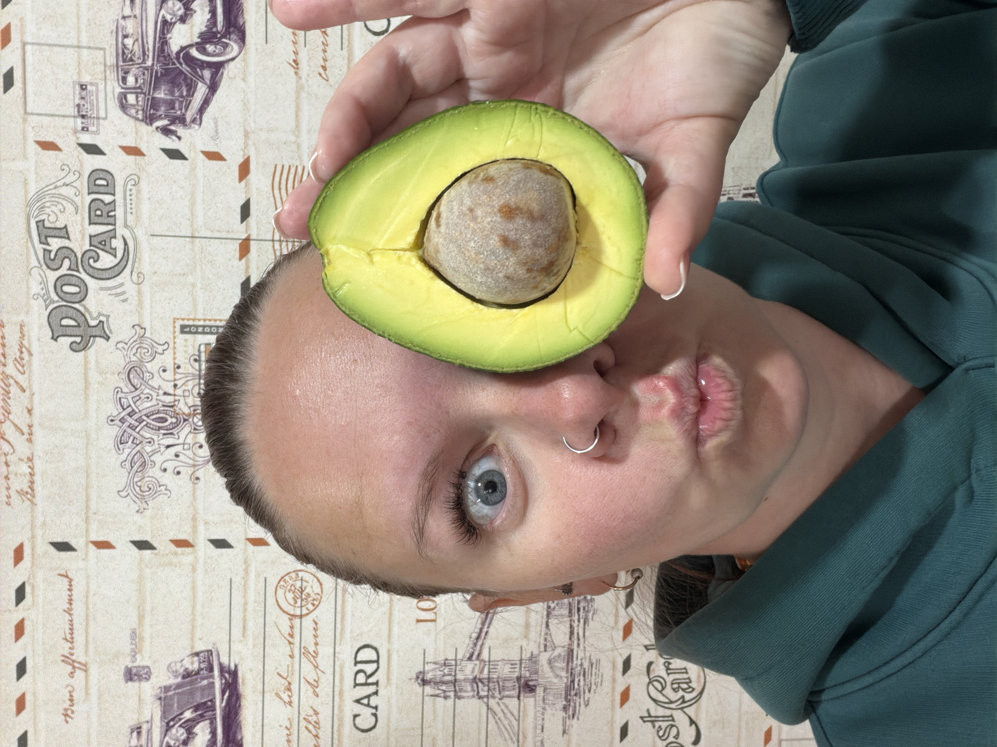 A humorous and playful portrait features a young person, located in Chaikovsky, Russia, holding half an avocado with its large pit over their right eye. The individual has fair skin, striking light blue eyes, and dark hair pulled back. A silver hoop nose ring is visible on their left nostril, and they are making a puckered-lip expression, contributing to the lighthearted mood of the image. They are wearing a dark teal or forest green hooded sweatshirt.
The image is oriented vertically, rotated 90 degrees clockwise, with the person's head positioned towards the lower left. The background is a light-colored surface, possibly a table or wall, adorned with a pattern resembling vintage postcards. These designs include illustrations of old cars and landmarks, specifically the Tower Bridge in London. Visible text fragments on the background include "POST CARD" (repeated), "LONDON," "GOLDEN," and partial or faded phrases such as "Bun afferturutement," "ne hier sauch," and "ebles de flemen." The setting appears to be indoors, under ambient lighting.