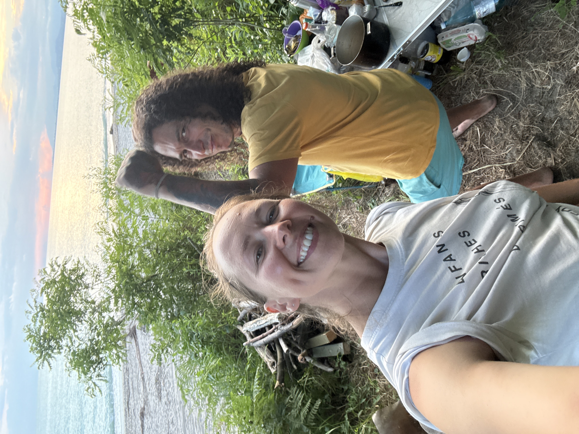 The image depicts a man and a woman in an outdoor setting near a body of water, likely a beach. The man, with long curly hair, is wearing a yellow t-shirt and blue shorts. He is flexing his bicep towards the camera. The woman, in the foreground, is wearing a white t-shirt with text on it and is smiling at the camera. Behind them, there is vegetation, a pile of firewood, and a table with various items, including cooking pots, bottles, and a carton of eggs. The background shows the sea and a distant shoreline under a cloudy sky with hints of sunset colors. The scene appears to be a casual camping or picnic setup. The location is identified as Yuada-Gulrypsh, Abkhazia.