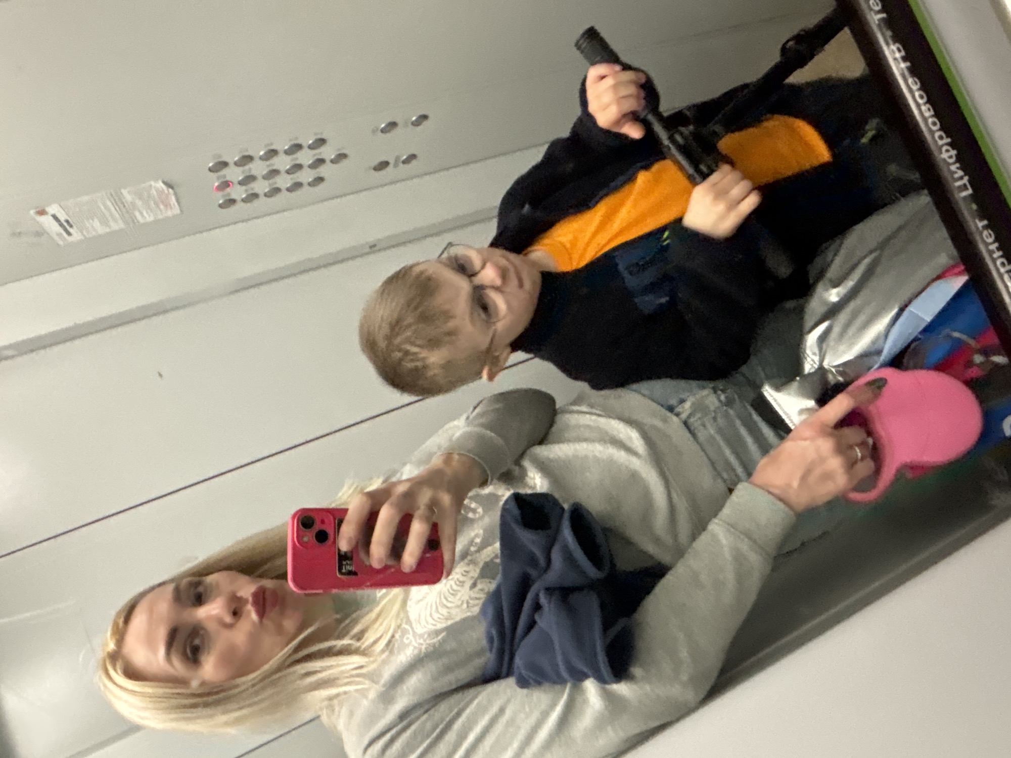 A selfie-style photograph shows a woman and a child inside an elevator. The woman, with blonde hair, holds a pink smartphone, presumably taking the picture. She wears a light grey sweatshirt and blue jeans. A blue article of clothing is visible on her lap. The child, wearing glasses, an orange shirt, and a dark jacket, is seated in a stroller. The child holds a dark, cylindrical object. A pink retractable dog leash is visible near the bottom right of the frame. Elevator buttons are visible on the wall above the woman. Writing in Russian can be seen in the upper right. Based on the Russian text "Лифтовое оборудование", the location is likely in Saint Petersburg, Russia.
