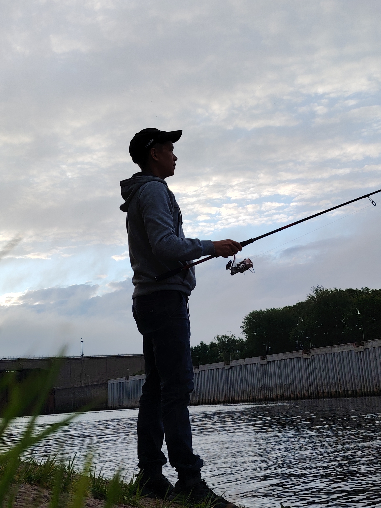 A full shot shows a person fishing on the bank of a river. The person, silhouetted against a cloudy sky, is wearing a dark baseball cap, a grey hooded sweatshirt, and dark pants. They are holding a fishing rod with their right hand, casting a line into the water. The river is calm, with small ripples on the surface. In the background, there is a concrete wall with vertical supports lining the riverbank, followed by a line of trees. A bridge or dam structure is visible further in the distance on the left. The scene appears to be an outdoor recreational area within the city of Chaikovsky, Russia, likely near the Kama River.