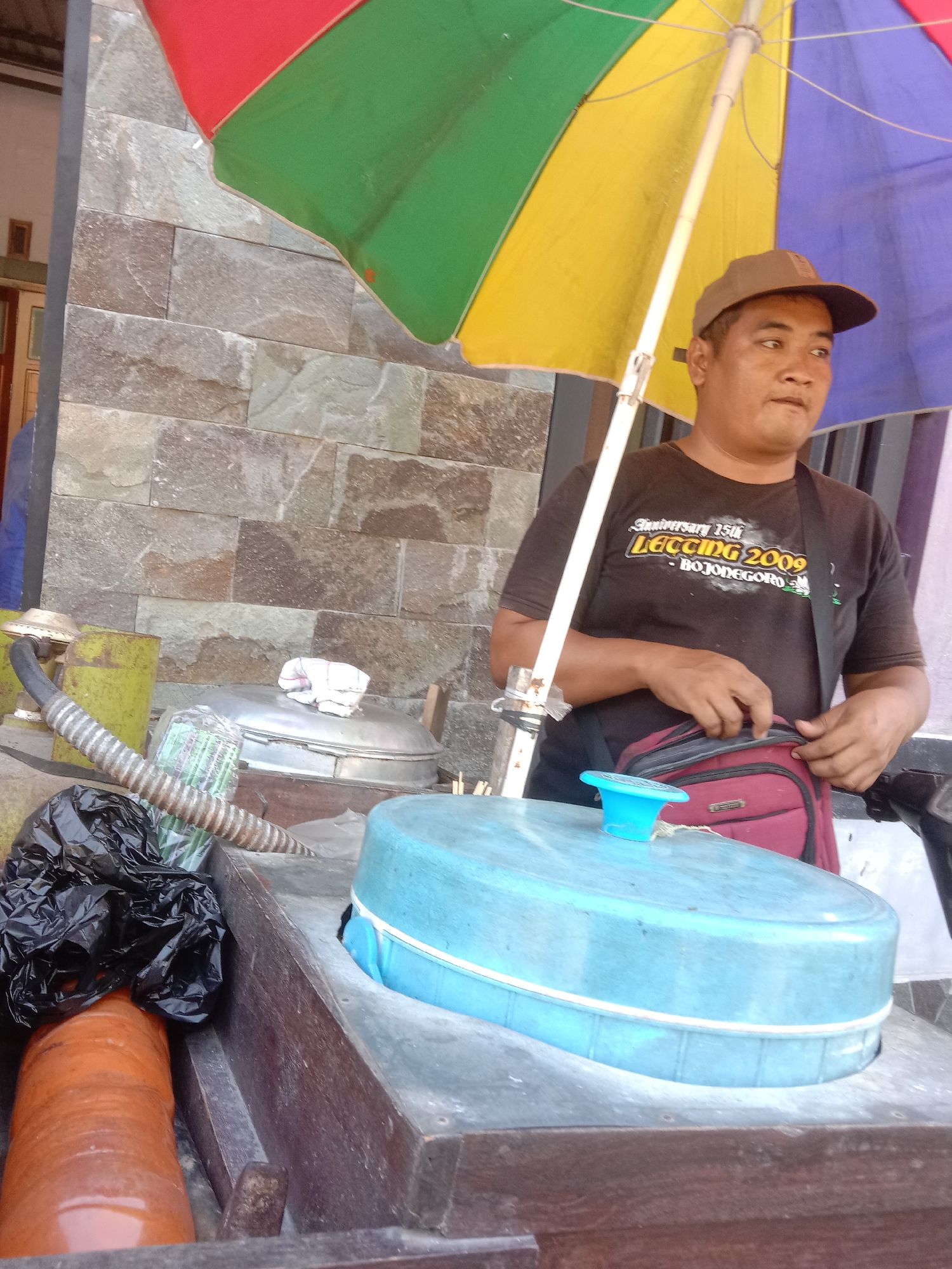 A man wearing a brown baseball cap and a dark t-shirt with the text "Anniversary 15th LEGUNG 2009 BOJONEGORO" is standing under a multi-colored umbrella. He is holding a maroon bag with his right hand. In front of him, on a wooden counter, are various objects including a large blue lid with a small blue funnel, a silver lid of a pot, a ribbed hose, a black plastic bag, and an orange cylindrical object. The background consists of a stone-tiled wall. The scene appears to be an outdoor stall, possibly a food vendor, in Tulungrejo, Indonesia.