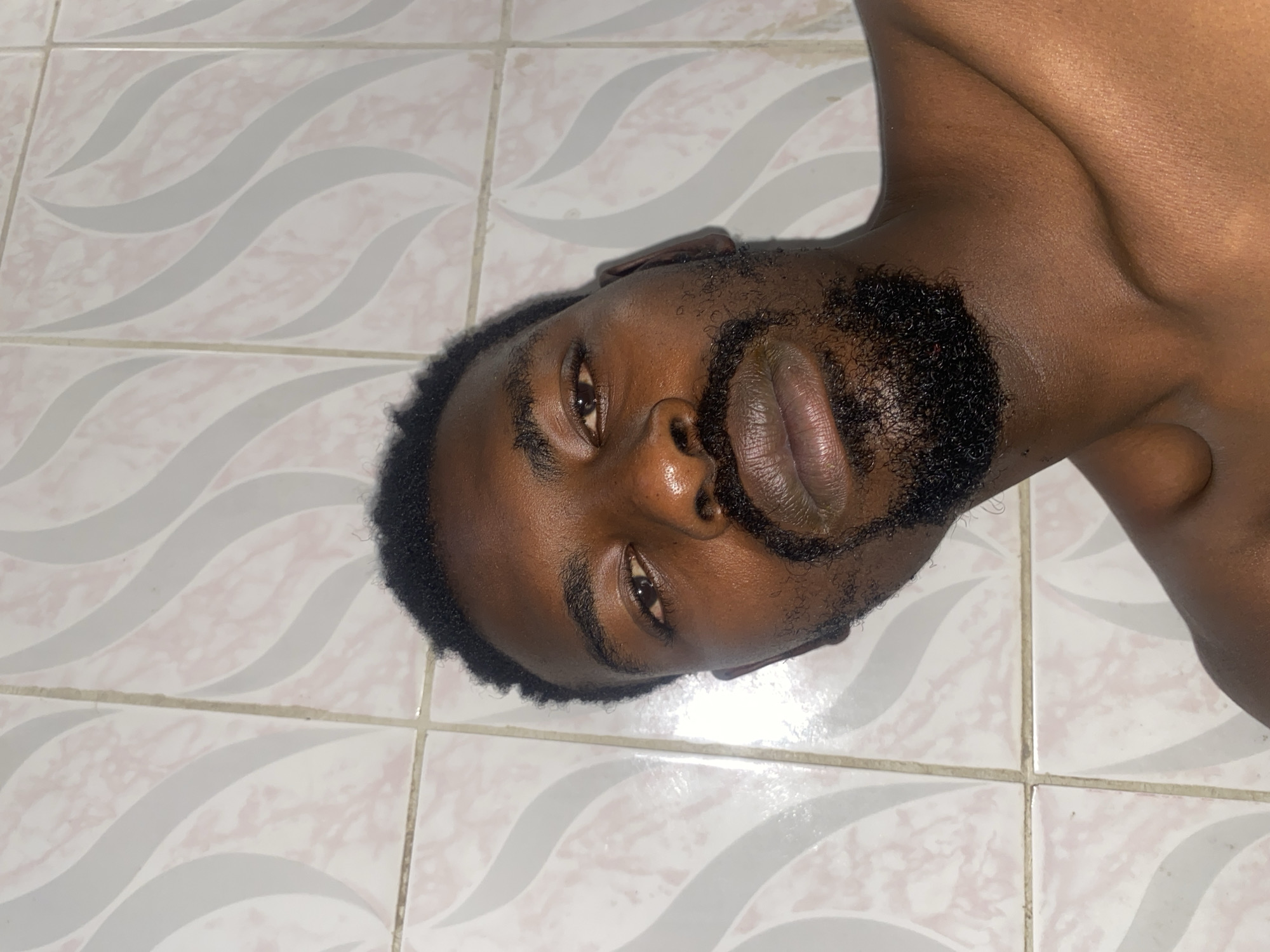 A dark-skinned man with a beard is lying on his side on a tile floor. The tile is white with a grey wavy pattern. He is shirtless. The exact location within Barakin Akawo, Nigeria is not discernible from the image.