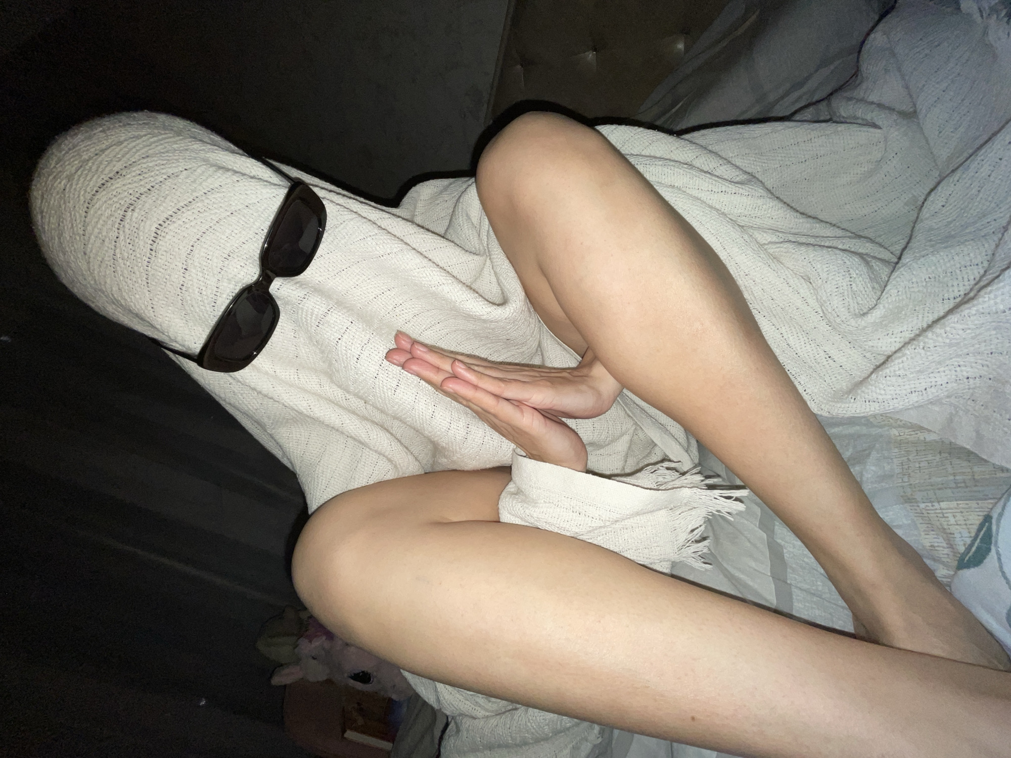 A person is lying in bed, draped in a light-colored, woven blanket. The blanket also covers their head, and a pair of dark sunglasses rests on the blanket-covered head. The person's hands are clasped together. Parts of the person's legs are visible. The background includes a headboard and what appears to be a stuffed animal and other objects near the bed. There's nothing in the image to confirm the location is Izhevsk, Russia.