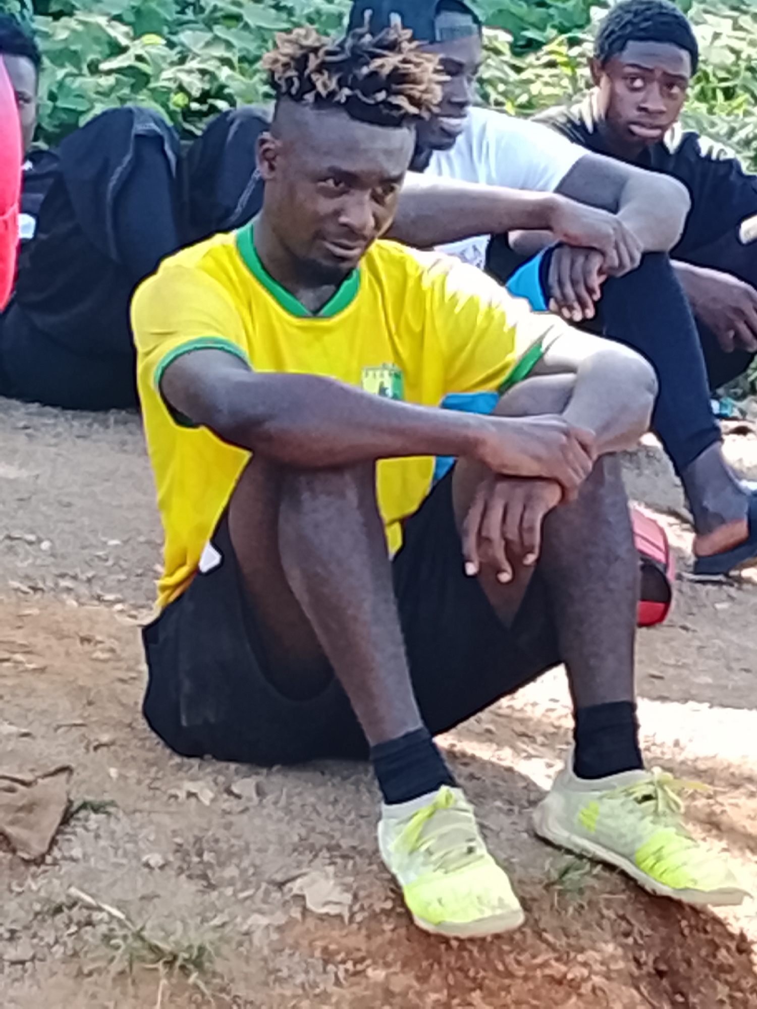 In Jos, Nigeria, three young men sit on the ground. The man in the foreground wears a yellow t-shirt with green trim and black shorts. His hair is styled in dreadlocks. To his left, another man, partially obscured, wears black clothing. To the right, a third man, also partially obscured, wears a white t-shirt and blue shorts, with a black baseball cap. The ground is composed of dirt and small stones. In the background, green foliage is visible. One man is wearing bright yellow athletic shoes.