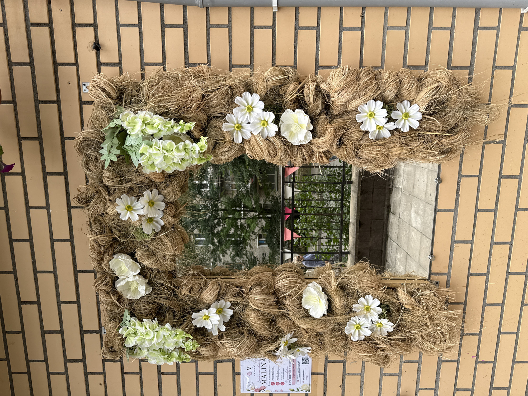 An archway made of coarse, brown twine is adorned with white and green artificial flowers. A mirror is mounted within the archway, reflecting a street scene with a brick building, trees, a metal fence, and a person walking. A poster with Cyrillic text and a logo featuring the letter "M" is attached to the left side of the archway. The archway is mounted on a light brown brick wall, with a gray downspout visible on the right. The scene appears to be outdoors, possibly in a public space in Gatchina, Russia, given the presence of the poster with Cyrillic text.