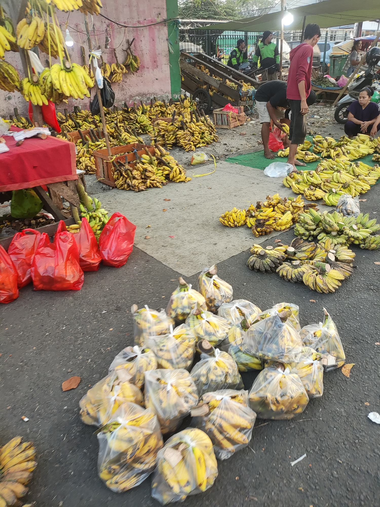 A street market scene in East Jakarta, Indonesia, features a large quantity of bananas displayed for sale. Ripe yellow bananas are prominently featured, both whole bunches hanging from a structure and laid out in piles. Some bananas are packaged in clear plastic bags, while others are in open wooden crates or directly on the ground. Red plastic bags are also visible, possibly for carrying purchases. Several individuals are present, some interacting with the produce, and others observing or engaged in other activities. A few motorbikes are parked in the background, suggesting a busy urban environment. The overall impression is of a vibrant, informal market setting.