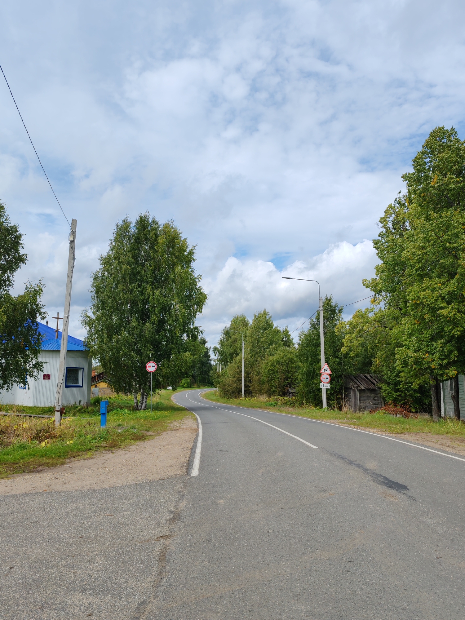 The image shows a rural road in Chukhlem, Russia, on a cloudy day. The road curves to the right, with trees lining both sides. On the left, there is a white building with a blue roof and a blue utility box on the side of the road. A traffic sign indicating a no entry for vehicles is visible near the building. Further down the road on the right, there is a weathered wooden shed and another traffic sign showing a winding road warning and a distance marker. The sky is filled with scattered white clouds against a light blue backdrop.