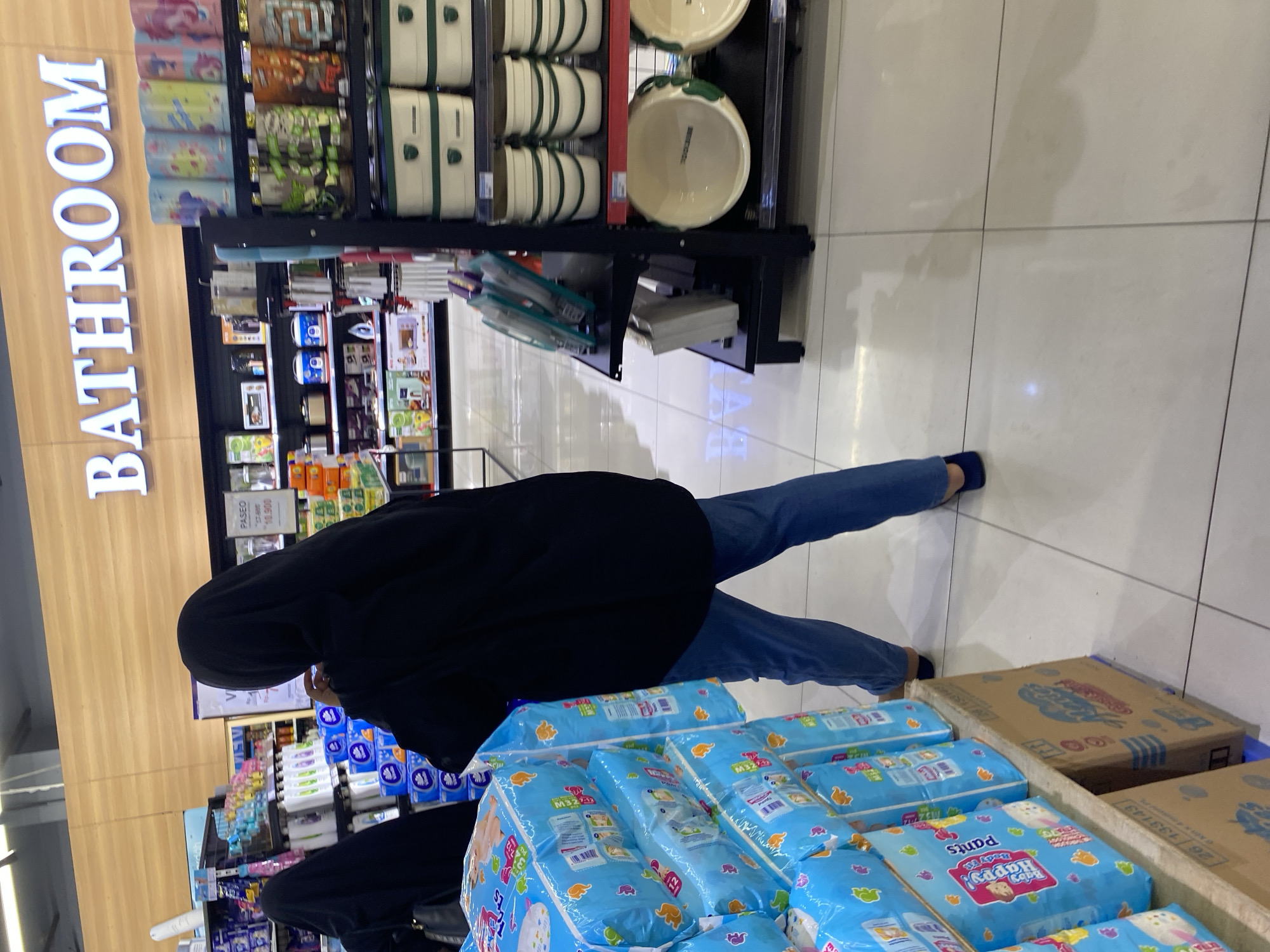 In a brightly lit supermarket in Banda Aceh, Indonesia, a person wearing a black hijab and black abaya with blue jeans walks past a display of baby diapers. The diapers are packaged in bright blue plastic with cartoon images. To the right of the diapers, a shelving unit displays various household items, including storage containers, bowls, and other bathroom-related products. Above the shelves, a large white sign reads "BATHROOM" in block letters. The floor is tiled with light-colored squares. Another person, also wearing a black hijab, is visible in the background to the left, partially obscured by the diaper display. The scene suggests a shopping trip for essential household goods.