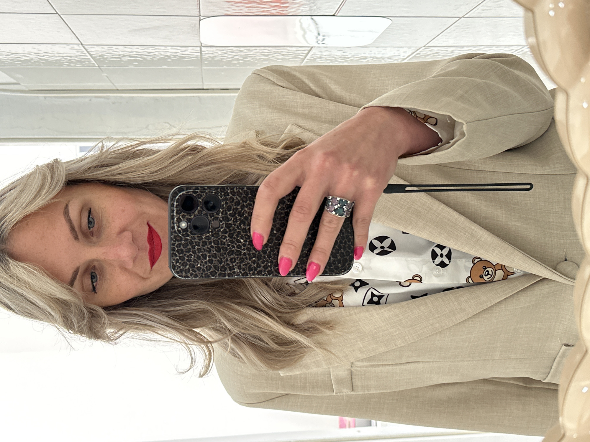 A blonde woman is taking a selfie in a mirror. She is wearing a beige blazer over a white patterned top. She has red lipstick and bright pink nail polish. She is wearing a large ring on her right hand. The reflection shows tiled walls. There is no evidence to indicate this scene is located in Venafro, Italy.