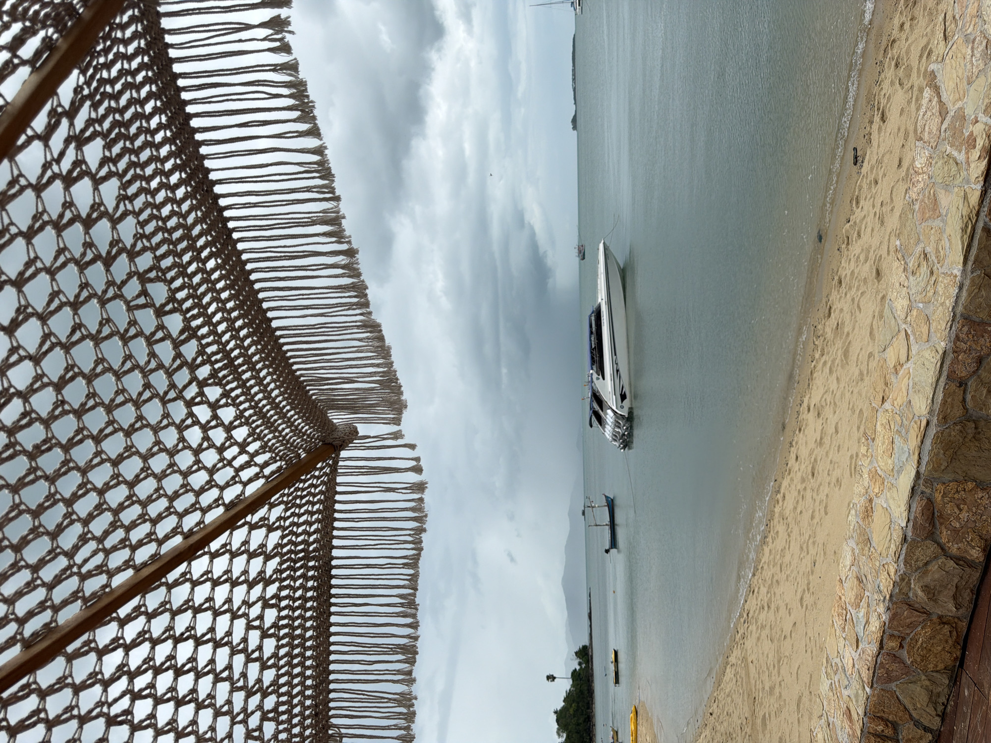 The image captures a coastal scene on a cloudy day, likely in the Ko Samui District of Thailand. In the foreground, a textured stone wall separates a sandy beach from a wooden deck. The beach slopes gently towards the calm, teal-colored ocean.
Several boats are visible in the water. The most prominent is a sleek, white speed boat with multiple engines, docked near the shore. Further out, a smaller, darker fishing boat with a blue canopy is anchored. In the distance, a sailboat can be seen. The water appears to be a tranquil bay.
The sky is heavily overcast with dark, dramatic clouds, suggesting the possibility of rain. Hazy mountains form the distant shoreline, with a single palm tree visible on the left side of the frame.
Overhead, a macrame-style umbrella or canopy, with a wooden frame and fringed tassels, partially obscures the view of the sky. The overall atmosphere is serene, with a touch of atmospheric drama due to the cloudy weather. No people are visible in the scene.