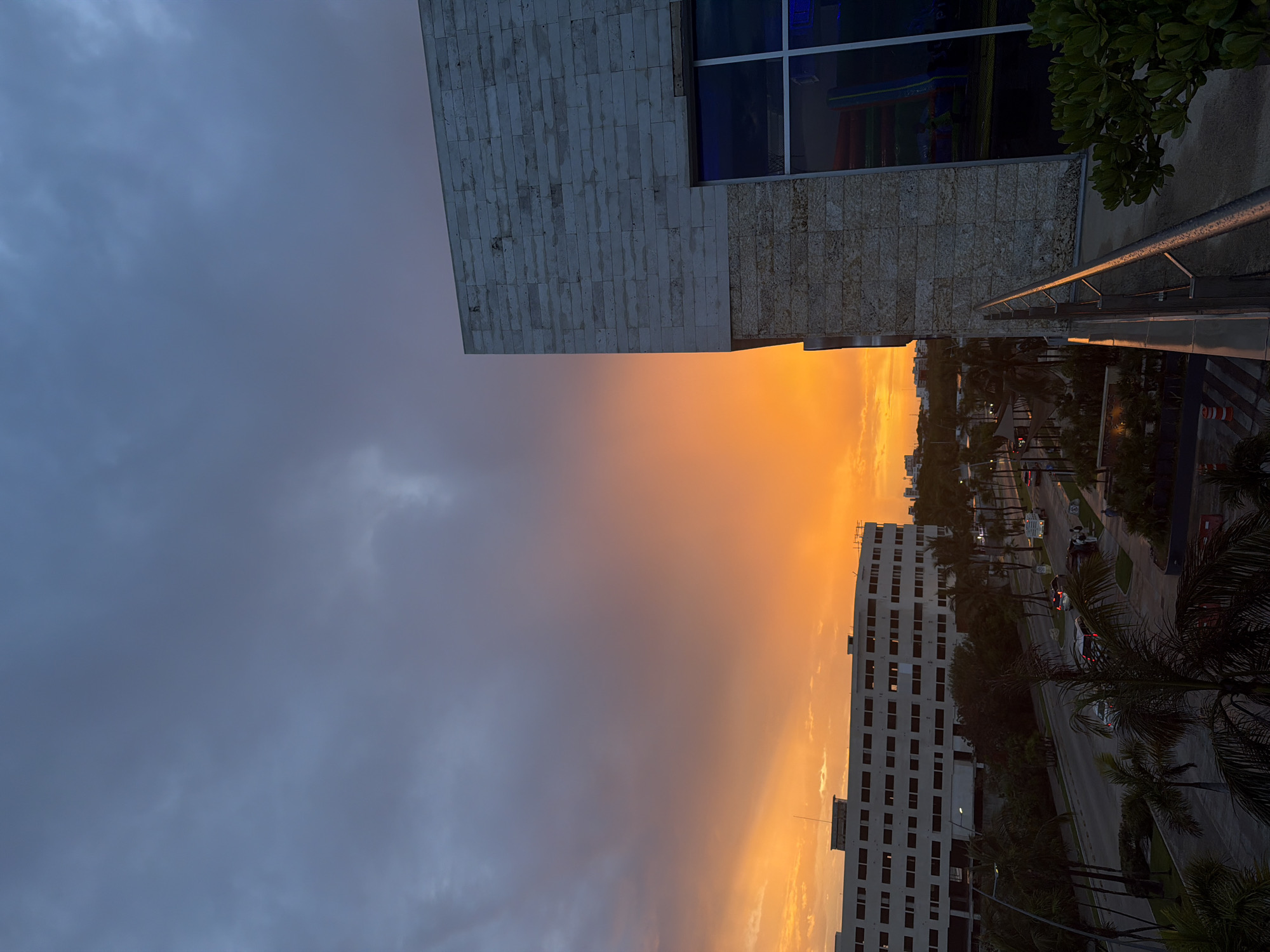 The image captures a dusky sky over Cancun, Mexico, with a vibrant orange and yellow sunset dominating the horizon. In the foreground, a modern building with a stone facade and large windows stands to the right. A balcony railing runs along the bottom right of the frame, with lush green foliage visible at the corner. To the left, a long, multi-story white building with numerous windows is situated across a street lined with palm trees and parked cars. The street appears to be a commercial or urban area, with traffic lights and pedestrian walkways visible. The overall scene suggests an urban sunset view from an elevated position, likely a hotel or residential building in Cancun.
