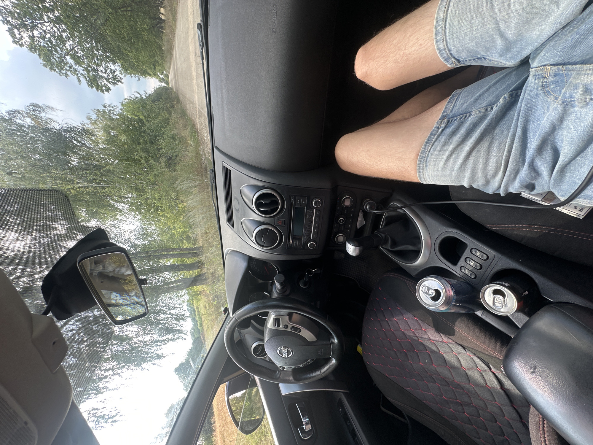 The image shows the interior of a car, likely a Nissan, with a view of a gravel road lined with trees and a field outside. The driver's legs are visible in the foreground, wearing denim shorts. Two cans of what appear to be soft drinks or beer are in the center console. The dashboard and steering wheel are visible, along with the rearview mirror. The car appears to be stationary on a dirt road in a rural area. The sky is cloudy. The specific location in Tynna, Ukraine cannot be determined from the image alone.