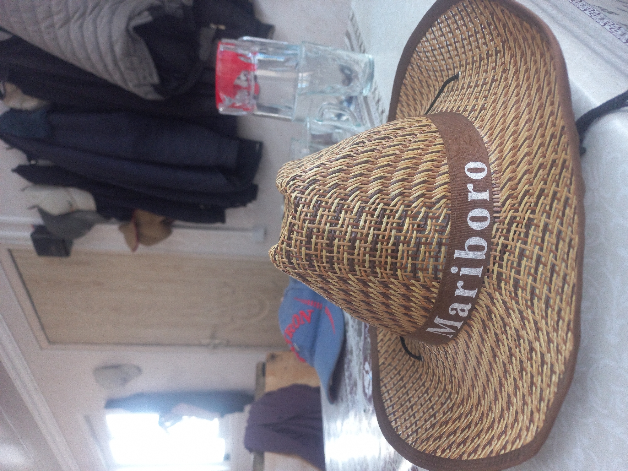 A straw cowboy hat with a brown band that reads "Mariboro" in white letters is the main subject of the image. The hat is placed on a table in the foreground. In the background, there is a blue baseball cap with red lettering, a purple garment draped over a chair, and a stack of clear glasses with a red-rimmed glass on top. Clothes hang on a rack behind the table. The scene appears to be indoors, possibly in a residential setting, with a window and a door visible in the background. The lighting suggests it is daytime. There are no people visible in the image. The location in the city of Şəfalı, Azerbaijan is not discernible from the image itself.