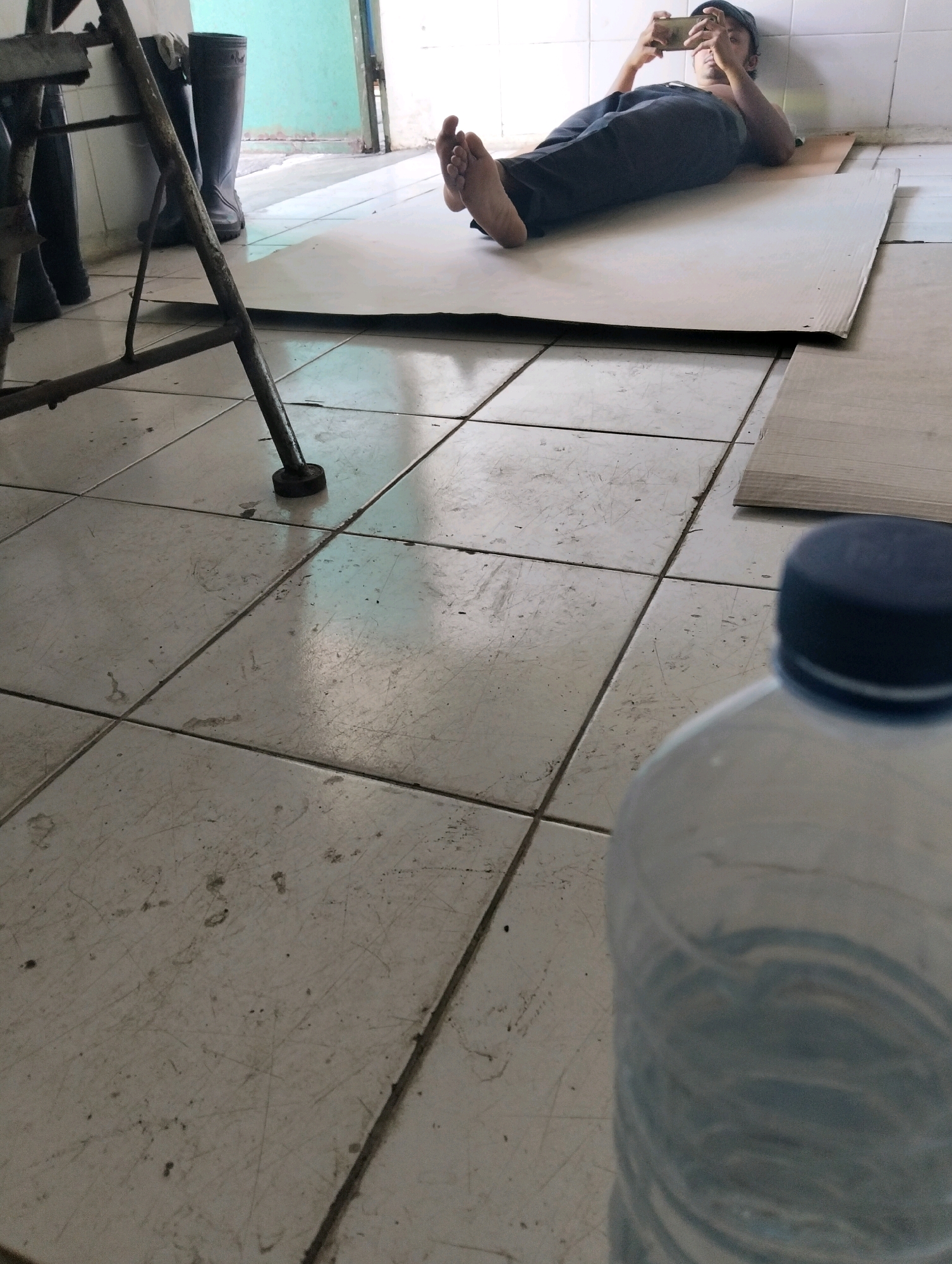 In a room with a tiled floor, a man lies on his back on a large piece of cardboard. He is wearing dark pants and a dark cap, and he is holding a smartphone in his hands, looking at the screen. His bare feet are propped up. To the left of the man, there are several pairs of dark rubber boots, and a metal structure that appears to be part of a chair or table. The background shows a wall and a doorway leading to an outdoor area. In the foreground, a plastic water bottle with a blue cap is partially visible. The overall scene suggests a casual, indoor setting.