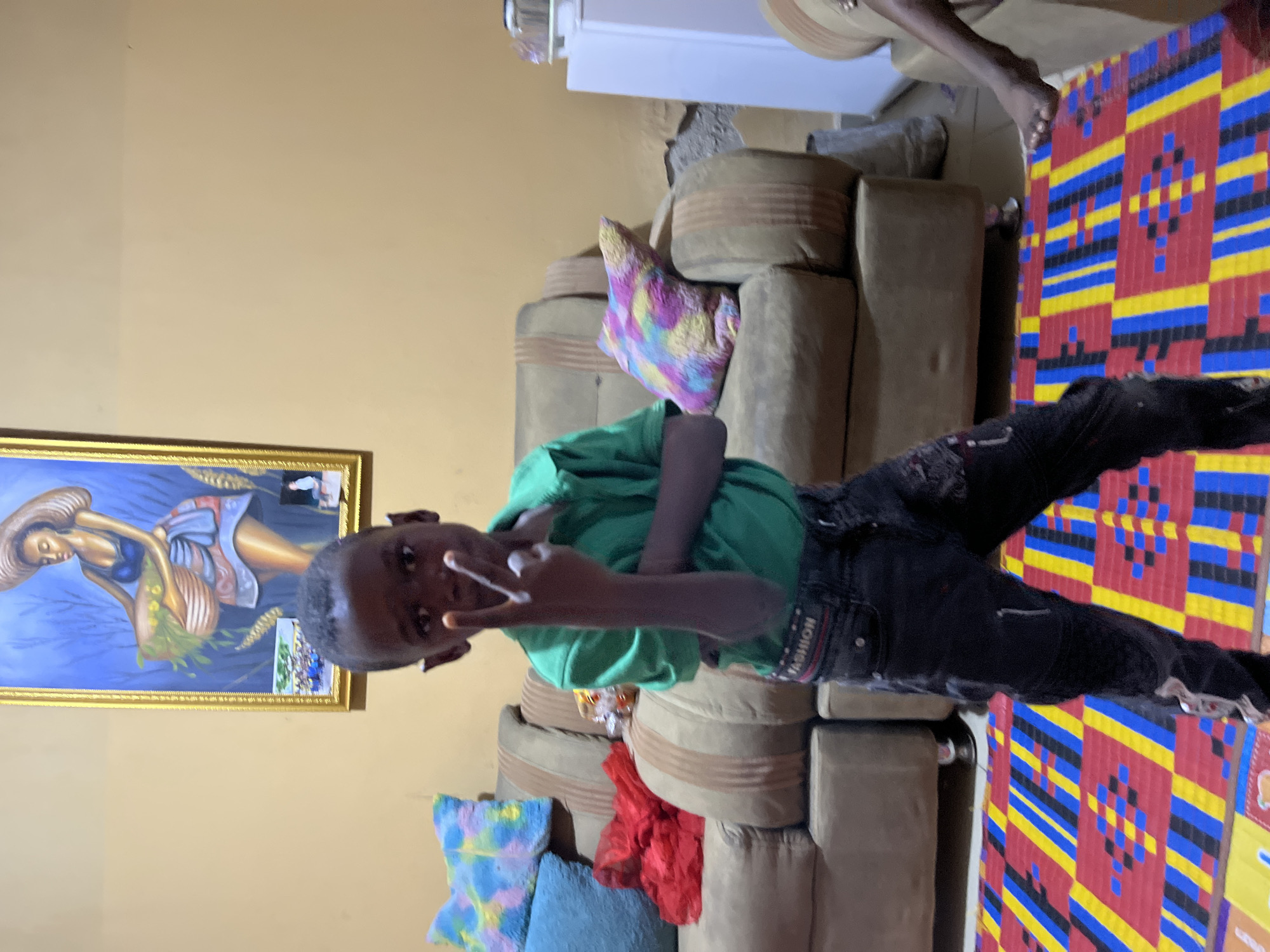 A young person is the primary subject in the scene, standing next to a brown sectional sofa. The individual is light-skinned, wearing a green shirt and black pants. They are making a V-sign with their fingers, and the overall impression is a casual, informal pose.
To the left is an artwork depicting a woman in a field, framed in gold. The background behind the couch and person is a solid, beige wall. On the sofa, various pillows are placed, exhibiting different colors and patterns. A patterned floor mat or rug with red, blue, black, and yellow motifs is on the floor.
The location is Madalla, Nigeria.
