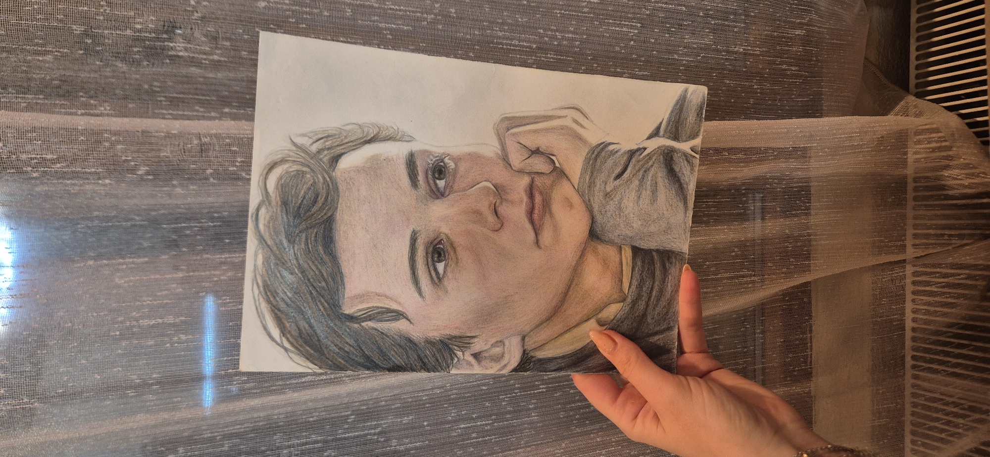 A medium shot depicts a hand holding a colored pencil drawing of a person. The drawing shows a person with dark hair, fair skin, and facial features rendered with shading and color. The hand holding the drawing has manicured fingernails and a gold bracelet. The background is a sheer curtain, partially illuminated by light coming through. A section of what appears to be a radiator or similar structure is visible behind the curtain. The location is indoors.