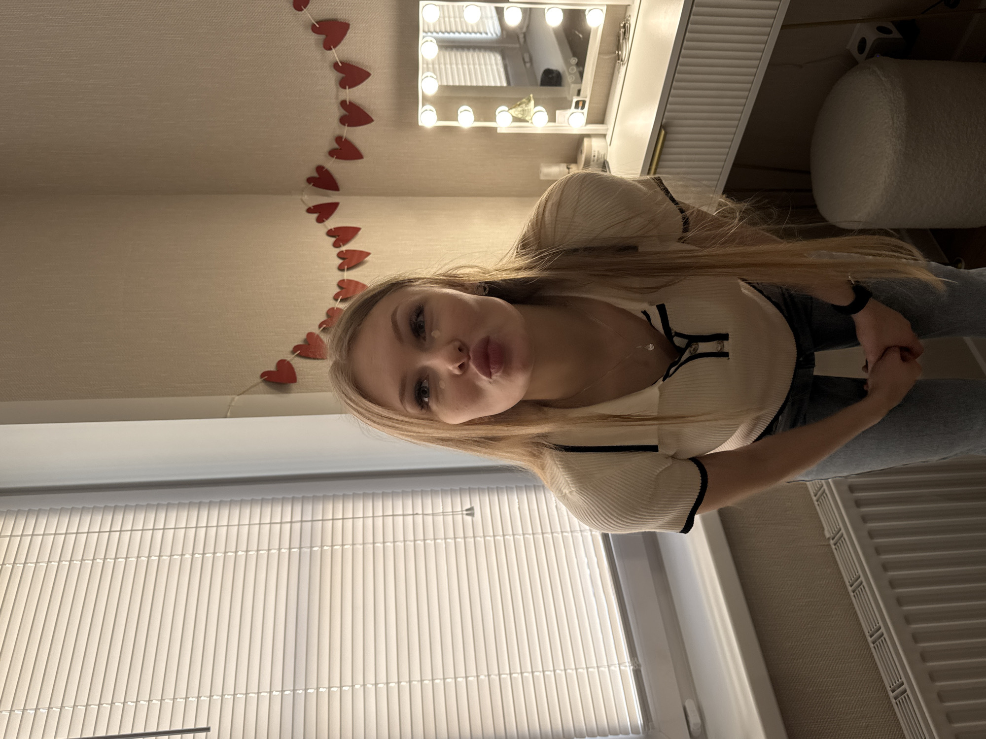 A blonde woman stands indoors in a room decorated with a string of red hearts, facing the camera and puckering her lips. She is wearing a white shirt with black trim and light-wash jeans. Behind her is a vanity with a light-up mirror, and a white stool is visible to the right. A window with blinds is present on the left side of the image. The location is identified as Perm, Russia.