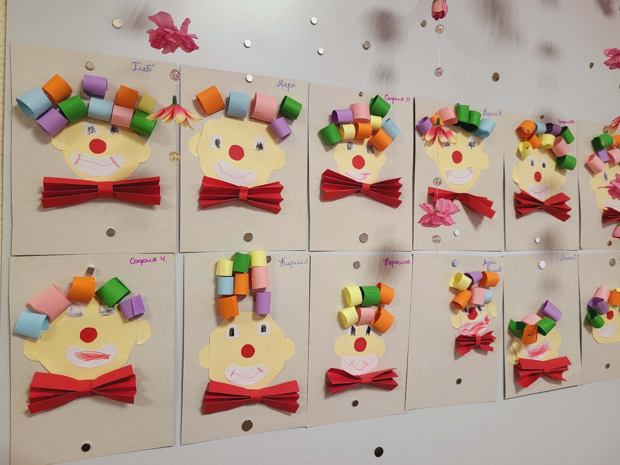 The image shows a collection of children's artwork displayed on a wall, likely in a kindergarten or preschool setting. The artwork consists of clown faces crafted from paper, with colorful curled paper strips representing hair and red paper bow ties. The artwork is pinned to the wall with what appear to be magnetic tacks. Each clown face is accompanied by a handwritten name in Cyrillic, indicating the artist. The names include "Глеб," "Марк," "София П," "Кириллл," "Софияя," "София 4," "Кириллл," "Вероника," and "Андрей Мочалов." Decorations, including pink flowers and beaded strands, are also present on the wall. The location is assumed to be in the city of Chaikovsky, Russia, based on the Cyrillic names.