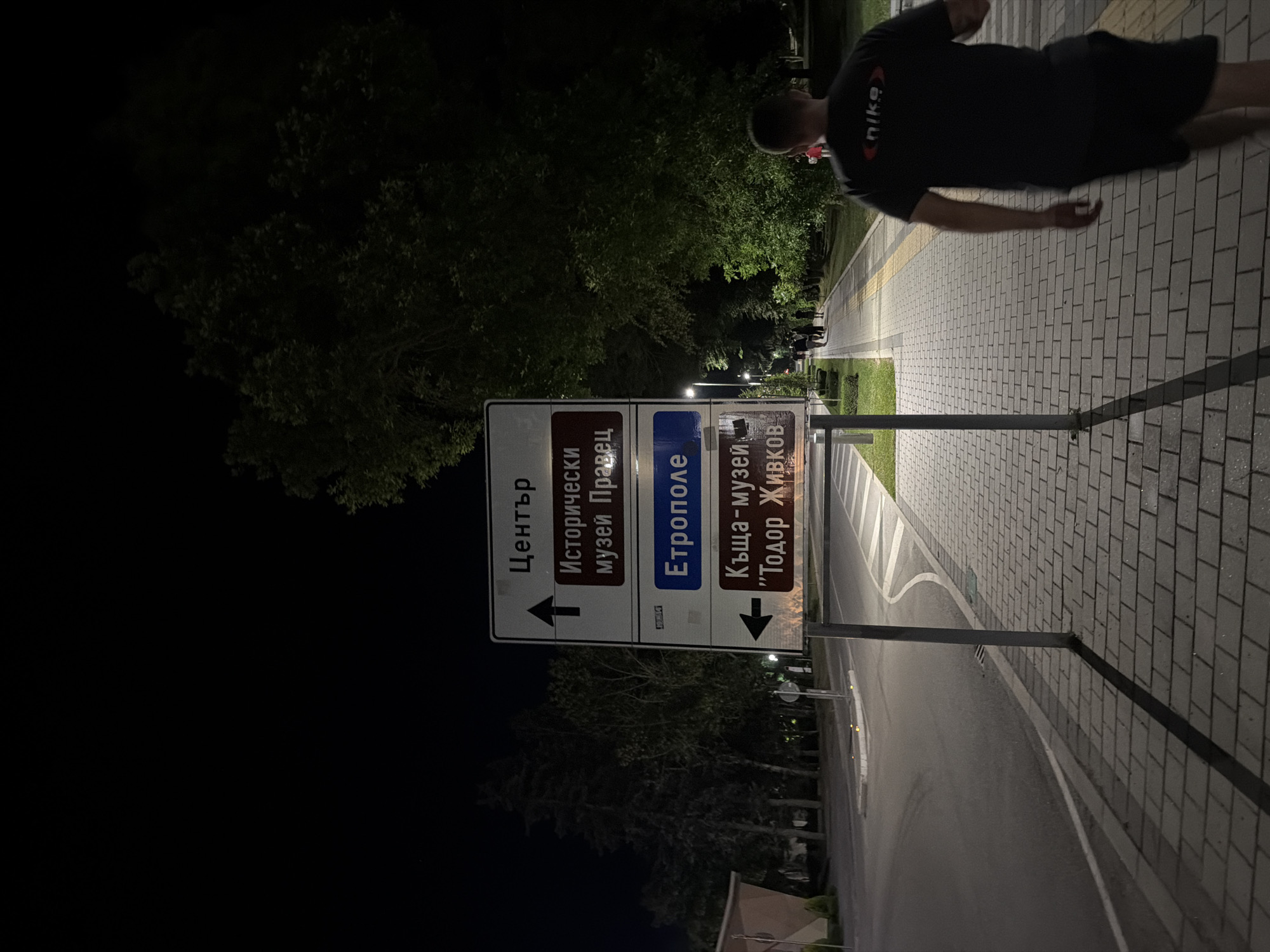 The image depicts a nighttime scene in the city of Pravets, Bulgaria, featuring a street, a sidewalk, trees, and a directional signpost. The signpost displays directions to "Център" (Center), "Исторически музей Правец" (Historical Museum Pravets), "Етрополе" (Etropole), and "Къща-музей 'Тодор Живков'" (House-Museum 'Todor Zhivkov'). A person is walking away from the camera on the sidewalk, wearing a dark t-shirt with a "Nike" logo. Streetlights illuminate the sidewalk and parts of the street. The environment appears to be a public area with pedestrian pathways and road infrastructure.