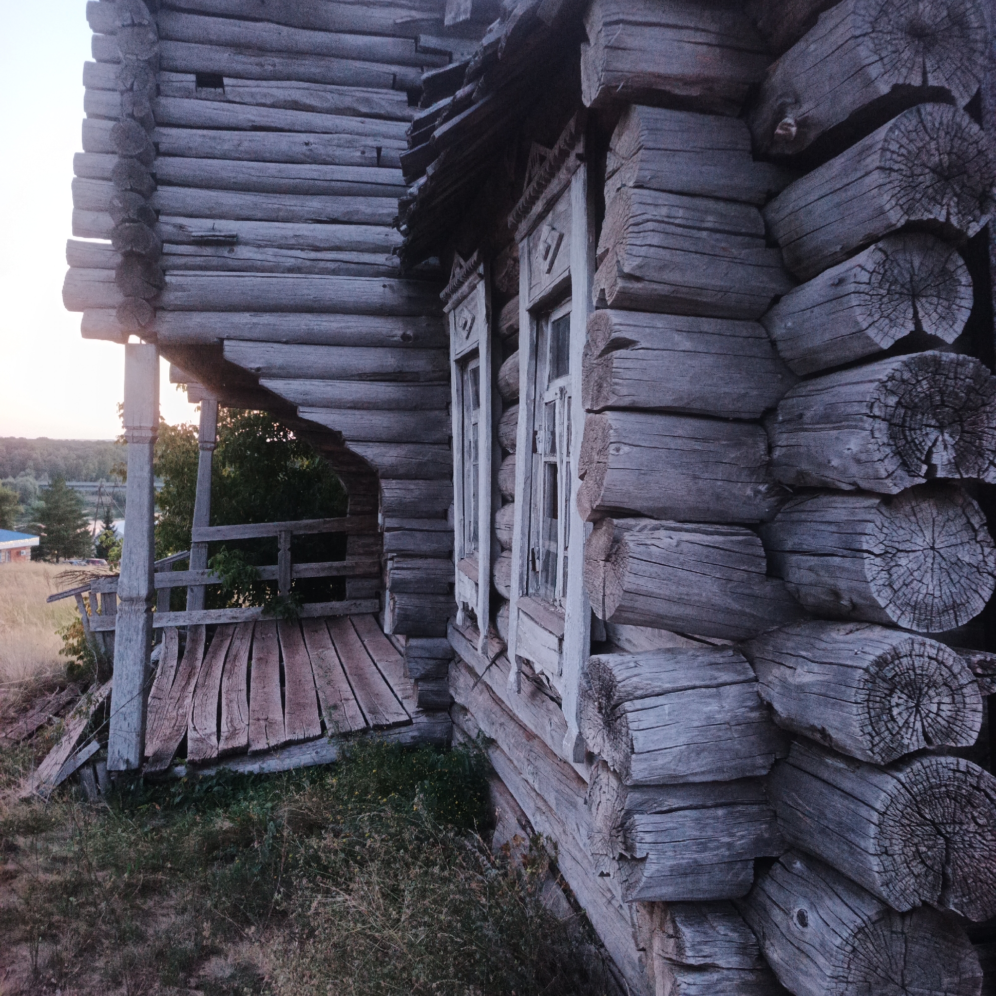 The image depicts the exterior of a weathered, unfinished wooden structure, likely a log cabin, situated outdoors. The building is constructed from large, rough-hewn logs, with visible grain and cracking, suggesting age and exposure to the elements. The architectural style appears rustic and traditional. To the left, a section of the building features an open porch with a wooden floor made of uneven planks, supported by thick wooden posts. The porch railing is also constructed of wood. Beyond the porch, the background reveals a landscape with dry grass, scattered trees, and a glimpse of what might be a body of water or a valley in the distance, under a pale, hazy sky, indicative of dawn or dusk. The overall impression is one of rural solitude and abandonment, possibly in a natural setting or on the outskirts of a village. The provided location, Tatarskii Saraktash, Russia, suggests this is a rural scene in that region. There are no people visible in the image.