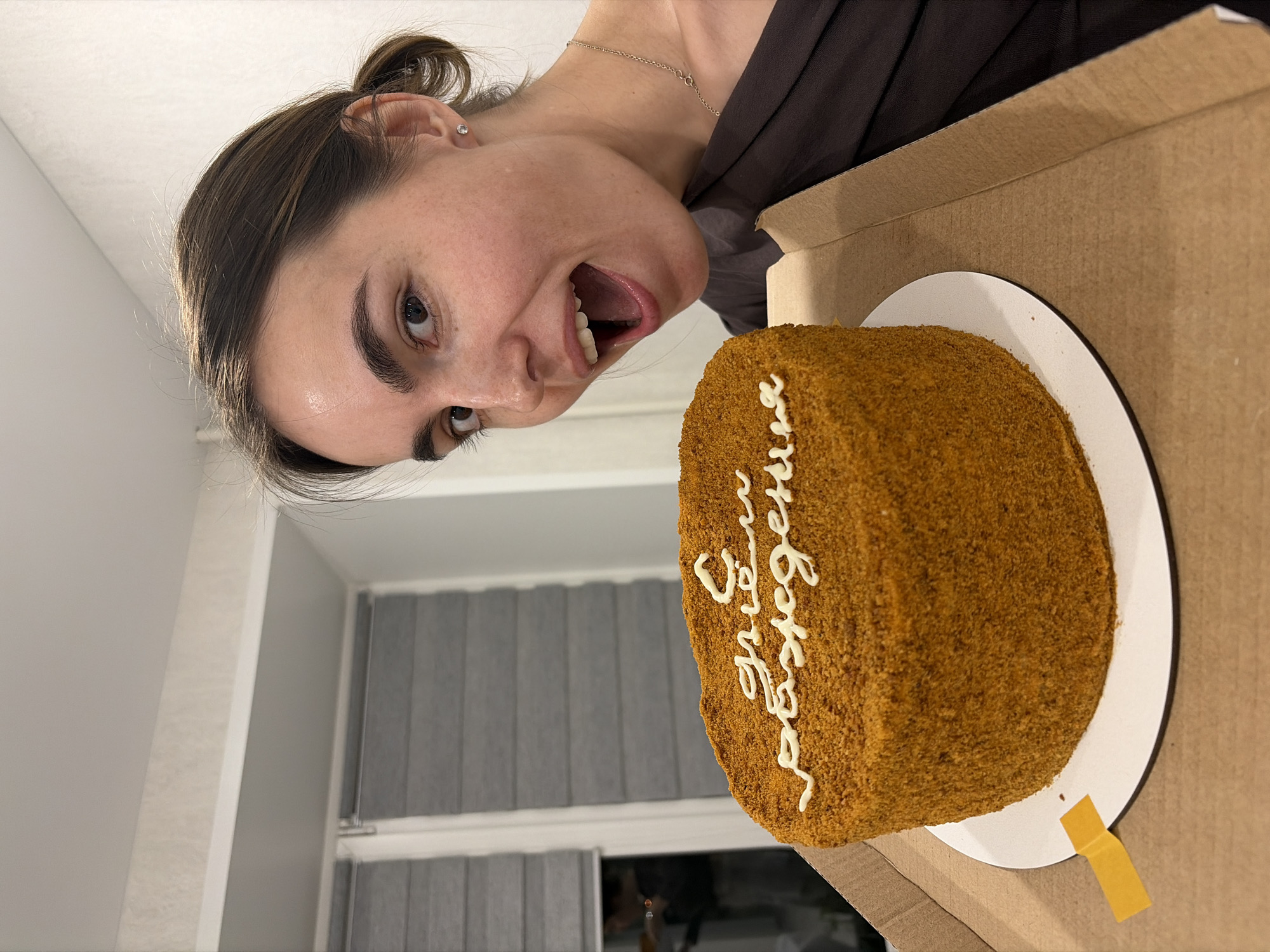 The image shows a woman with brown hair pulled into a bun, positioned to the left of a brown cake. The cake is on a white plate inside a cardboard box. White icing decorates the cake with Cyrillic text. The woman is tilting her head towards the cake with her mouth open. Behind the woman and to the left, a window with partially drawn blinds is visible. The image was taken in Sarapul, Russia.
