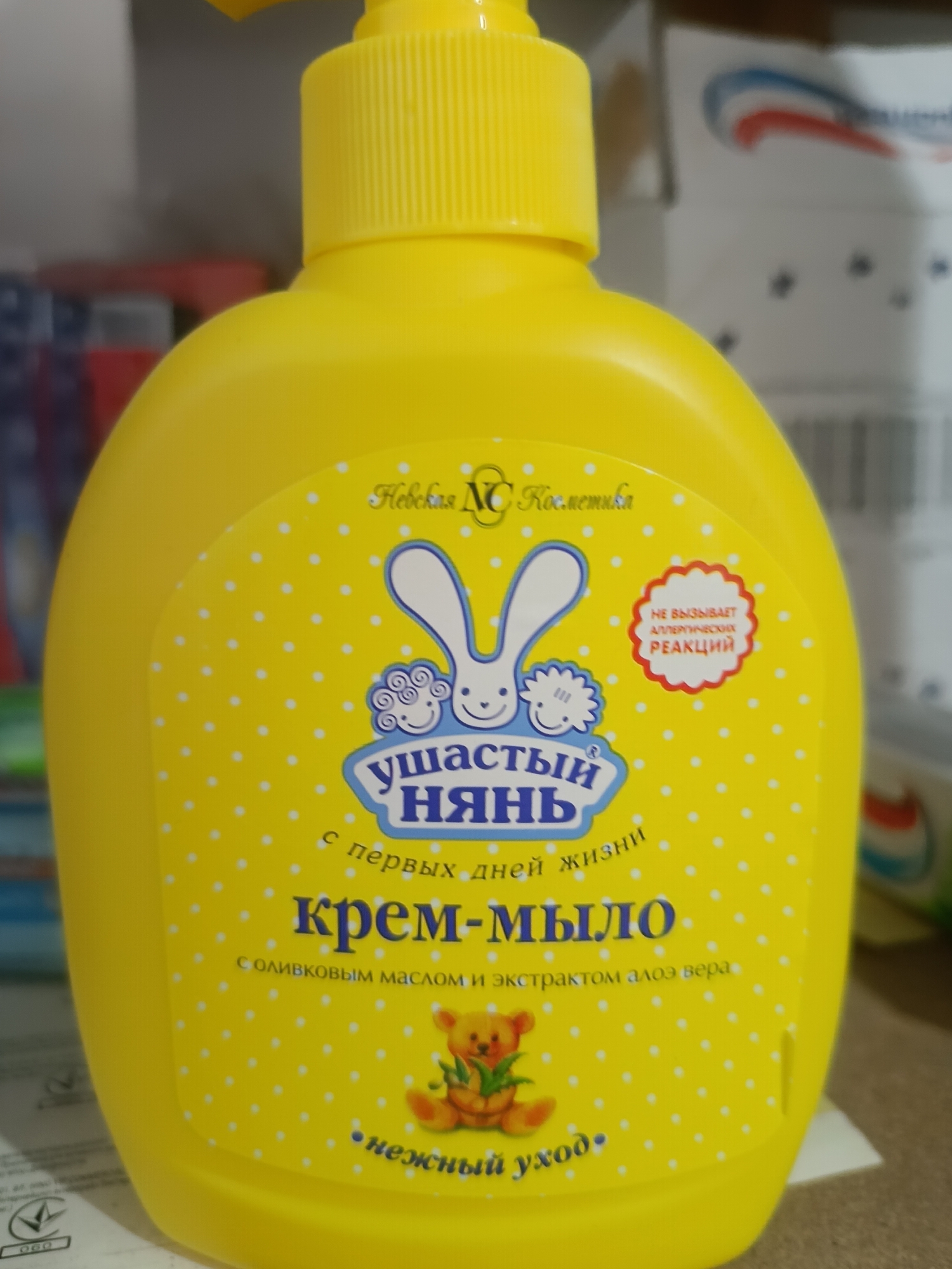 This media file features a bright yellow plastic bottle of "Ushastiy Nyan" (Ушастый Нянь) cream soap, designed for babies. The bottle has a pump dispenser at the top. The label is yellow with white polka dots and features illustrations of a cartoon bunny and a teddy bear.
Prominently displayed on the label are several lines of text in Russian. At the top, it reads "Невская NC Косметика" (Nevskaya NC Cosmetics). Below that, in a blue banner, is the brand name "Ушастый Нянь" (Ushastiy Nyan). Underneath, it states "с первых дней жизни" (from the first days of life). The main product name, "крем-мыло" (cream soap), is written in large blue letters. Further text below mentions "с оливковым маслом и экстрактом алоэ вера" (with olive oil and aloe vera extract). At the bottom, the phrase "нежный уход" (gentle care) is visible.
To the right of the bunny illustration, a red, scalloped circular badge contains text in Russian that translates to "DOES NOT CAUSE ALLERGIC REACTIONS". The overall impression is of a gentle, child-friendly product. The background is blurred but appears to be a retail setting with shelves and other products, suggesting the item is for sale. There are no discernible cues for time of day or weather. The location context indicates Berdiansk, Ukraine.