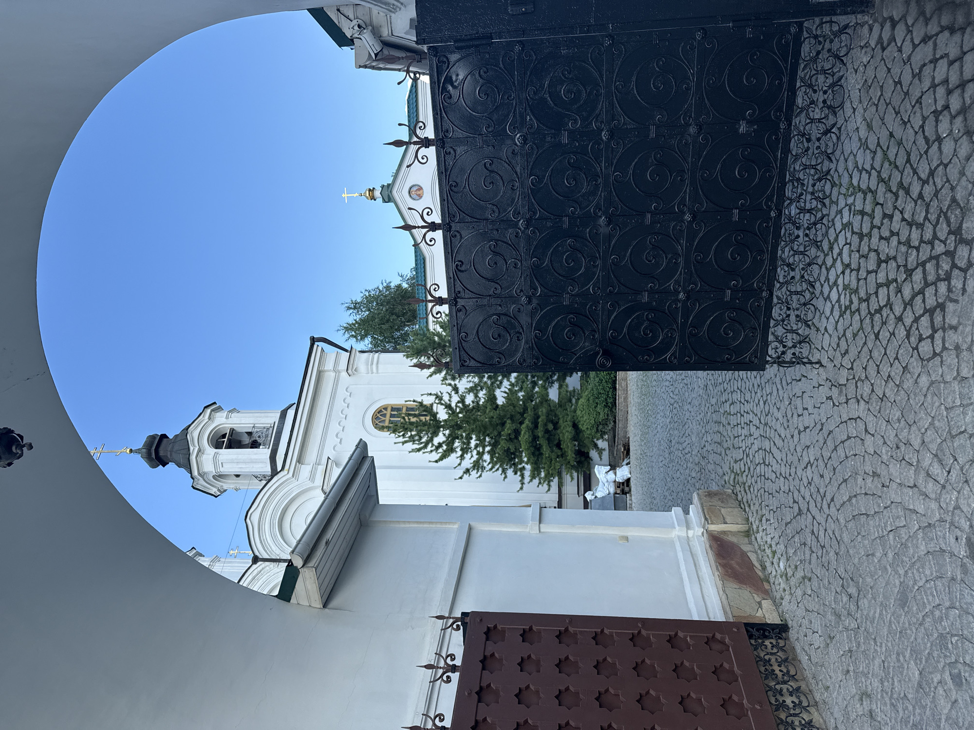 The image shows the entrance to a church complex in Yekaterinburg, Russia. The view is through a large white archway, framing a view of a white church with a black bell tower and green domes, under a clear blue sky. In the foreground, there are two gates: a red metal gate on the left and a large black wrought-iron gate on the right, which is partially open. The ground is paved with cobblestones. A small evergreen tree is visible to the left of the church, and a white statue of a seated figure can be seen near the tree. The overall scene suggests a peaceful and serene religious site.