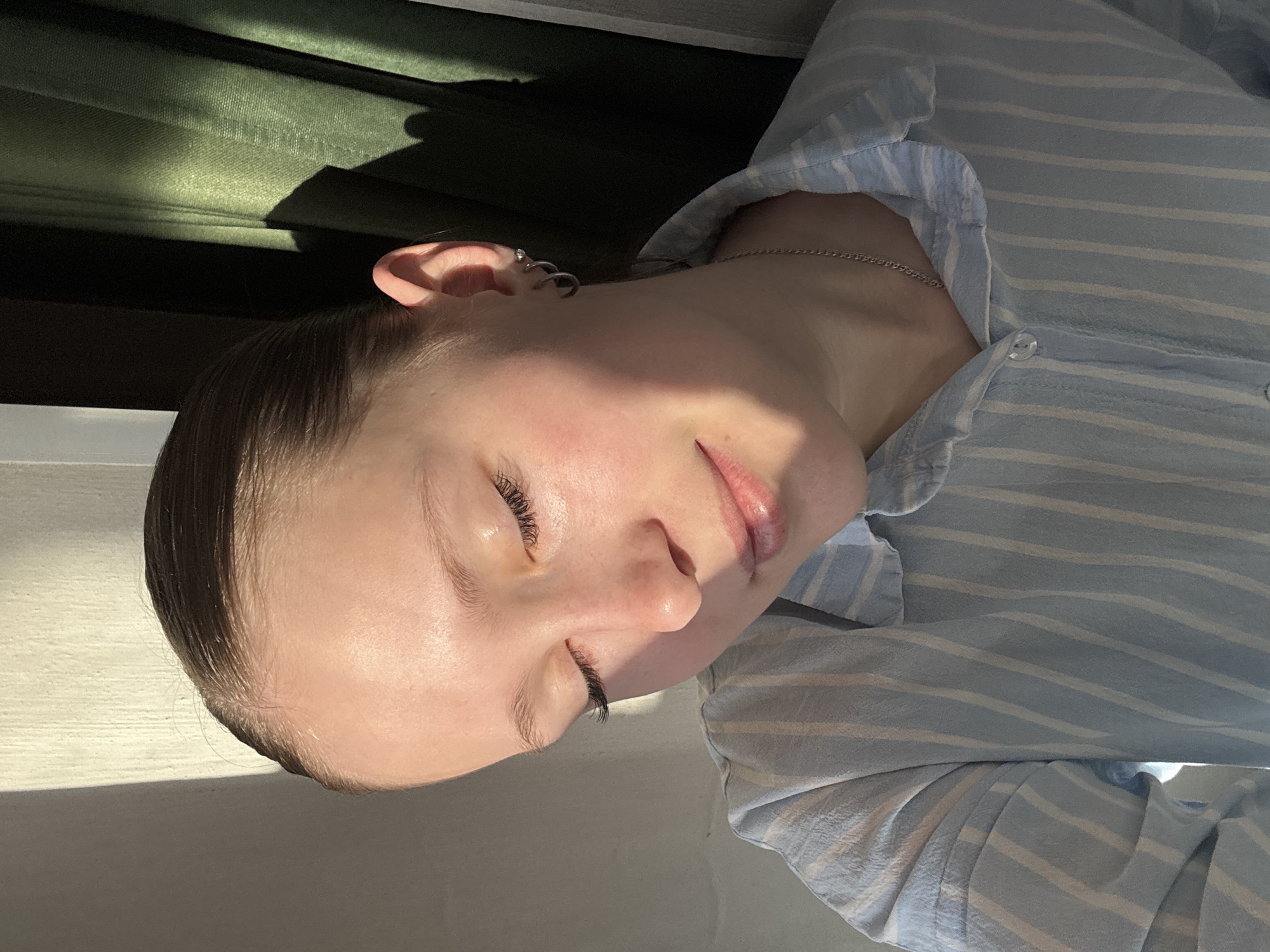 A close-up portrait captures a person, likely a young female, in a state of repose, bathed in direct sunlight. The image is oriented with the person's head to the left, turned slightly away from the viewer. They have fair skin, their eyes are gently closed, and a subtle, serene smile graces their lips, suggesting peace or relaxation. Long, dark eyelashes are prominent against their skin.
The individual wears a light blue and white striped button-up shirt made of a textured fabric. A thin silver chain necklace is visible around their neck, and their left ear is adorned with small silver hoop and stud earrings.
Strong, natural sunlight illuminates the scene, casting distinct highlights and sharp shadows across the face and shirt, indicative of a bright morning or late afternoon. The light originates from the top-left of the frame. In the upper part of the background, a dark green, textured fabric, possibly a curtain or blind, is visible, partially in shadow. A lighter, featureless surface is seen towards the bottom-left. The setting appears to be indoors, near a window, in Chaikovsky, Russia. There is no visible text.