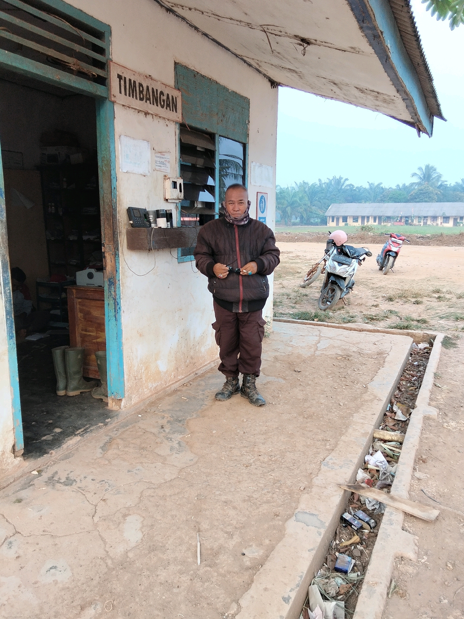 A man stands in front of a building with a sign that reads "Timbangan" in Indonesian, likely indicating a weigh station or scalehouse. The building appears weathered, with peeling paint and a corrugated metal roof. To the left of the building, an open doorway reveals shelves stocked with items and a desk. Green rubber boots are neatly placed near the doorway. The man is wearing a dark jacket, brown pants, and sturdy boots. He is holding a pair of glasses. In the background, several motorcycles are parked, and a long, low building with a series of windows can be seen. Palm trees and other vegetation suggest a tropical or subtropical environment. The ground is dirt and concrete, with a visible drainage ditch filled with debris. The sky is overcast. The location is identified as Teluk Tenggirik, Indonesia.