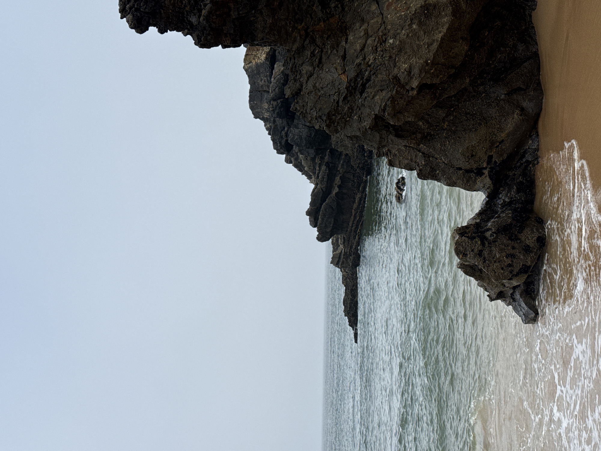 This is a vertical, eye-level shot of a rocky coastline on an overcast day. In the foreground, a sandy beach meets the calm, choppy ocean water. The water is a murky green-blue and is beginning to recede, leaving a foamy white tide line. To the right, a large, dark, jagged rock formation juts out towards the sea, dominating the right side of the frame. Further to the left, smaller rock formations break the surface of the water and extend out towards the horizon. The sky is a uniform, pale gray, with no visible sun. The scene is located in the Praia da Adraga, near Colares, Portugal.