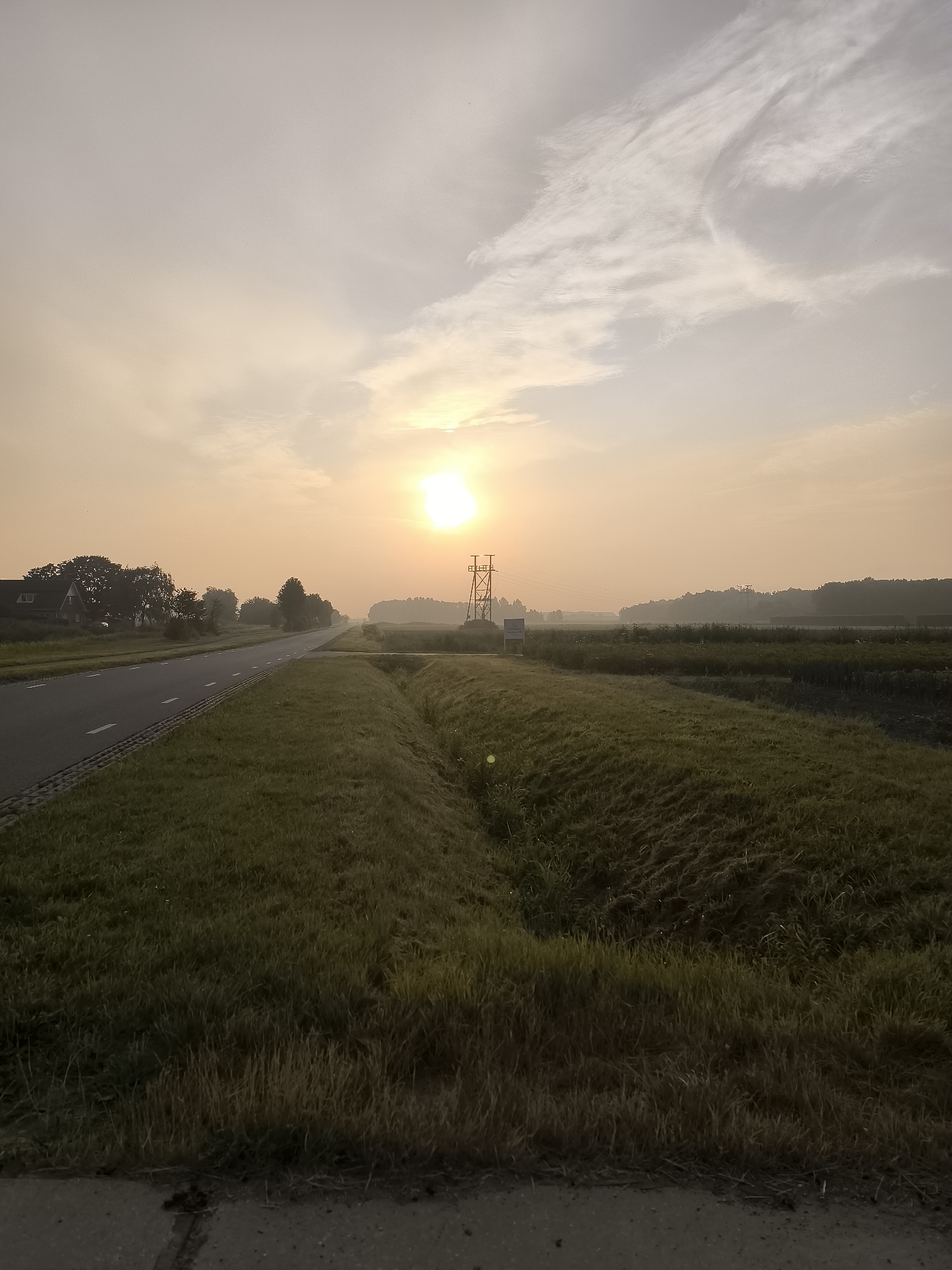 The image depicts a rural road in Andijk, Netherlands, during a sunrise. A paved road runs diagonally from the lower left to the middle of the frame, bordered by a grassy verge. The verge transitions into a field of vegetation with visible rows, suggesting agriculture. In the background, a line of trees and distant buildings are partially obscured by mist or haze, common in the early morning in this region. The sun, a bright orb, is visible in the upper right quadrant of the sky, casting a warm, diffused light. A tall, lattice-like structure, possibly a pylon or a wind turbine component, stands near the road in the mid-ground. The sky is a soft, peachy hue with wispy clouds. The overall scene evokes a tranquil, early morning atmosphere.