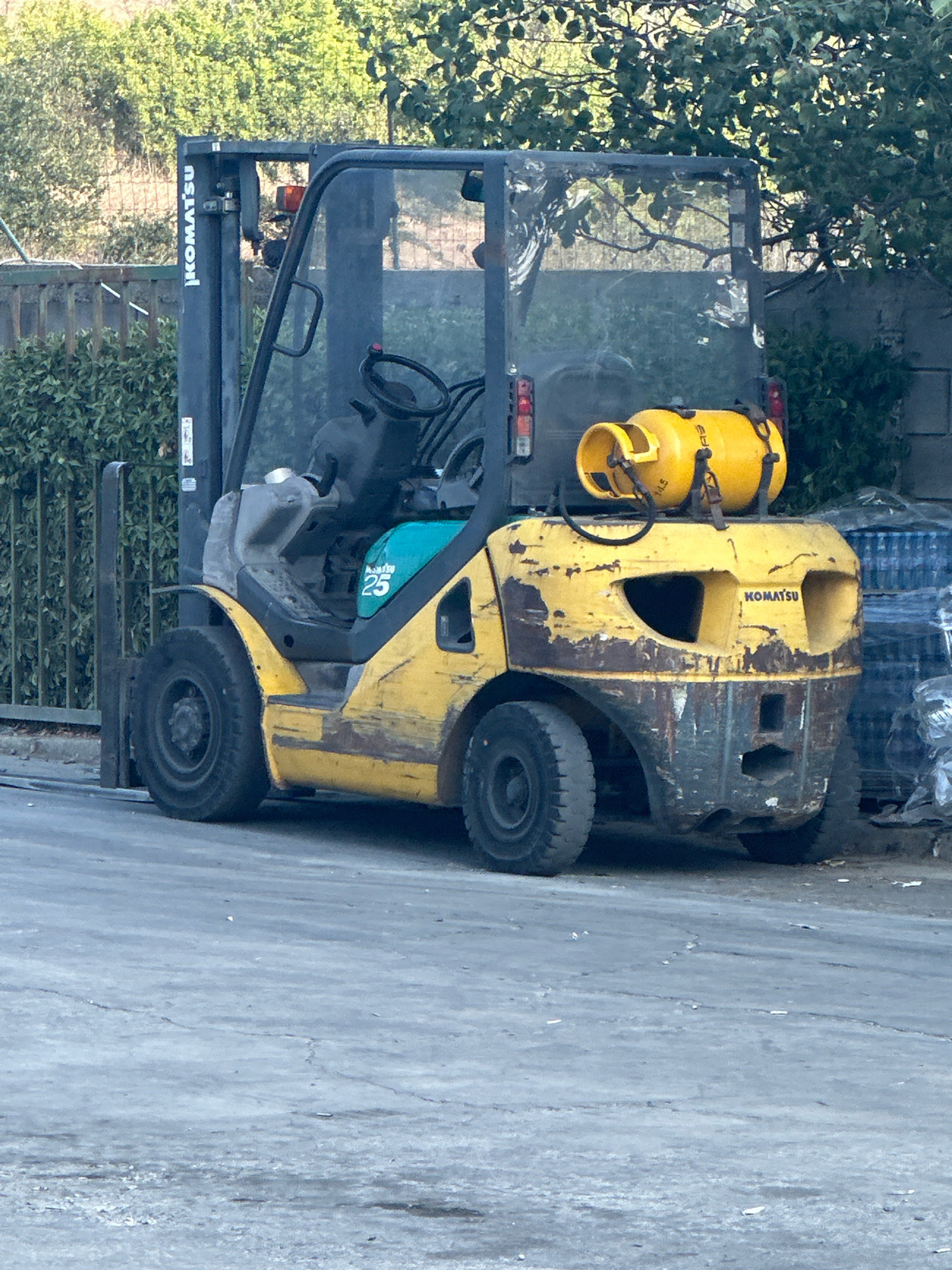A yellow and gray Komatsu forklift is parked on a cracked asphalt surface. The forklift is weathered, with rust and chipped paint visible on its body. A yellow propane tank is secured to the top rear of the forklift. The forklift is positioned in front of a green metal fence and dense green foliage. In the background, there are trees and a gray wall. The scene is outdoors, under daylight. The location is the city of Ravadinovo, Bulgaria.