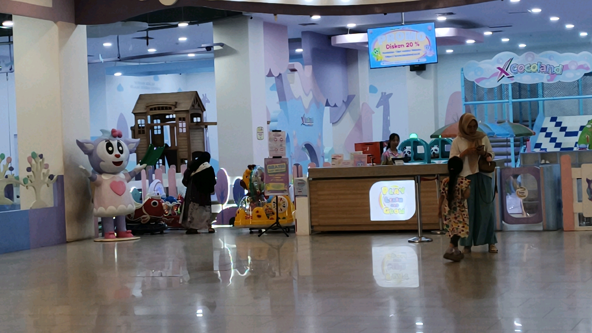This media file captures an indoor scene at "X Cocoland," a children's play area, likely a mall attraction in Baturaja, Indonesia. The brightly lit space is decorated with soft pastel colors and playful imagery, including cartoon giraffes and clouds. In the foreground, a large purple sheep mascot with a pink bow and dress stands near a small wooden playhouse with a slide. To the right, a wooden counter serves as a reception area, with signs visible that advertise "PROMO Diskon 20%".
Several people are present, including two women and two young girls. One woman, wearing a hijab and a white top with blue pants, appears to be checking her phone while standing near the counter, with a young girl in a colorful dress facing away from her. Another woman, also in a hijab, is partially visible near the playhouse. Two individuals, presumably staff members, are behind the counter, one of whom is interacting with a customer.
A small yellow toy ride is also visible, adding to the playful atmosphere. The floor is highly reflective, mirroring the lights and the objects in the scene. The overall impression is that of a lively, family-friendly entertainment venue designed for children. The time of day is indeterminate, but the indoor setting suggests it could be any time, likely during operating hours for the play area.