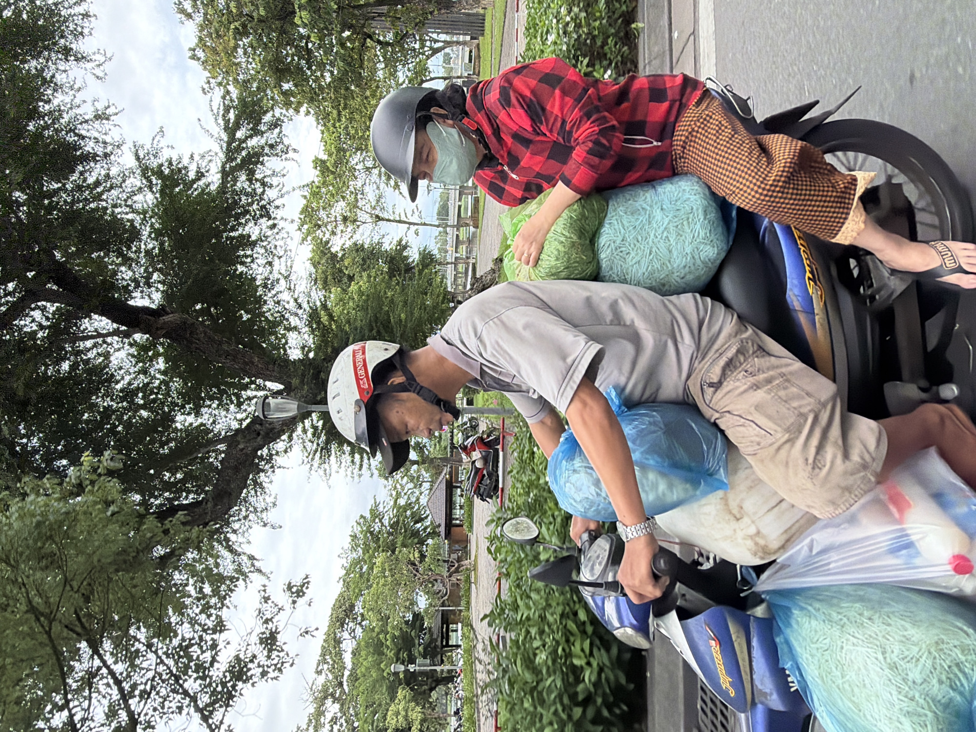 A man and a woman are riding on a blue motorcycle. The man is driving and wearing a grey shirt, khaki shorts, and a white helmet with red accents. The woman is sitting behind him, wearing a red and black checkered jacket, checkered pants, and a black helmet. She is also wearing a light green face mask and holding a large green bag. The motorcycle is carrying several large plastic bags filled with what appears to be bundled goods. The scene is set in Thuận Hóa Ward, Vietnam, with lush green trees and foliage visible in the background. The sky is overcast. The motorcycle is on a paved road with a curb and some greenery to the right. In the distance, there are more trees, buildings, and a bridge. The overall impression is of a busy urban scene, possibly during a delivery or transport of goods.