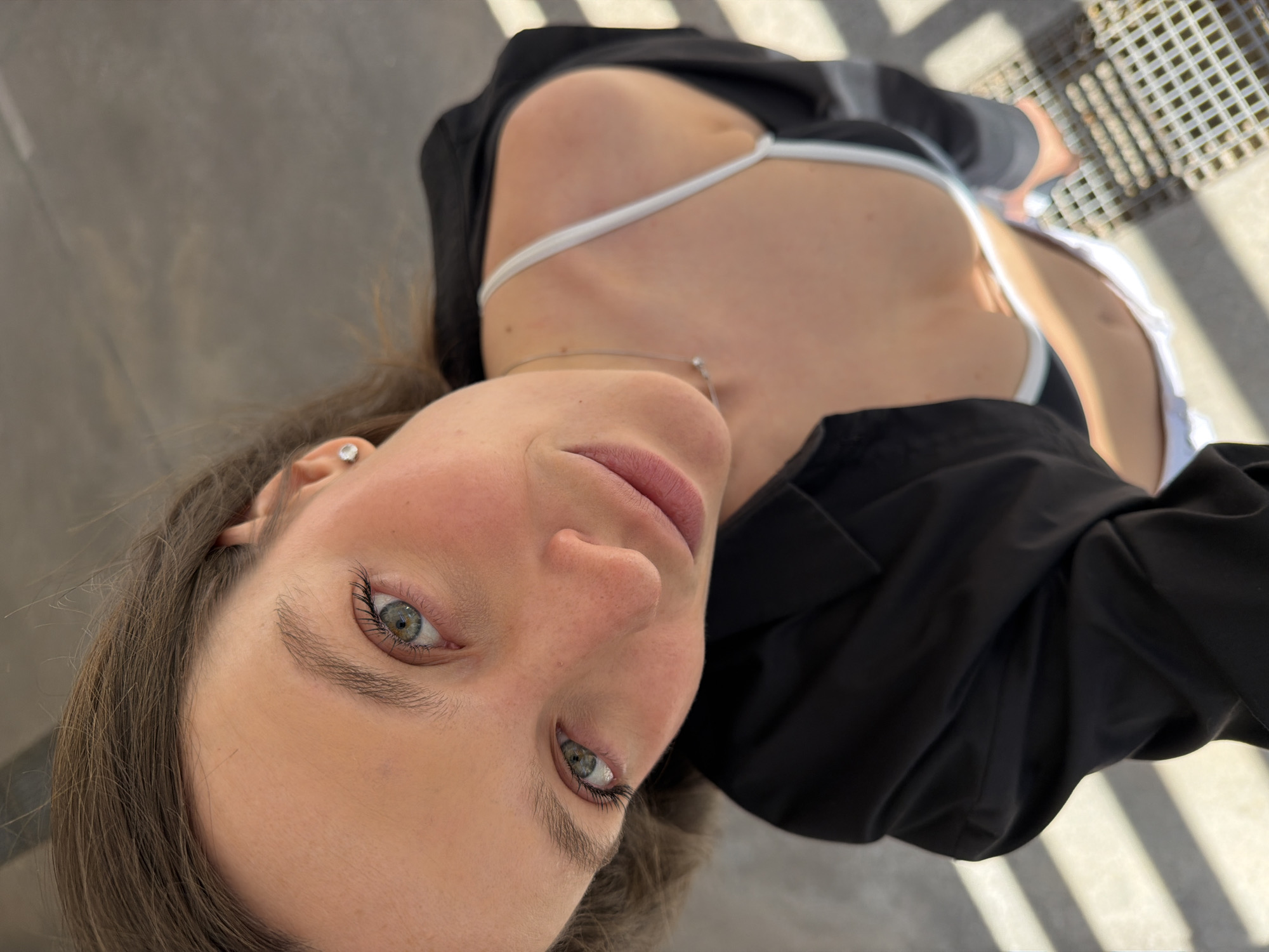 A low-angle, selfie-style photograph depicts a woman wearing a black jacket, a white swimsuit, and a silver necklace. The photograph's background shows a textured, gray surface and a metal grate with light patterns. The woman has fair skin, light brown hair, and blue eyes. A small earring is visible in her left ear. The photograph's location, Budva, Montenegro, is not identifiable from the visual elements.